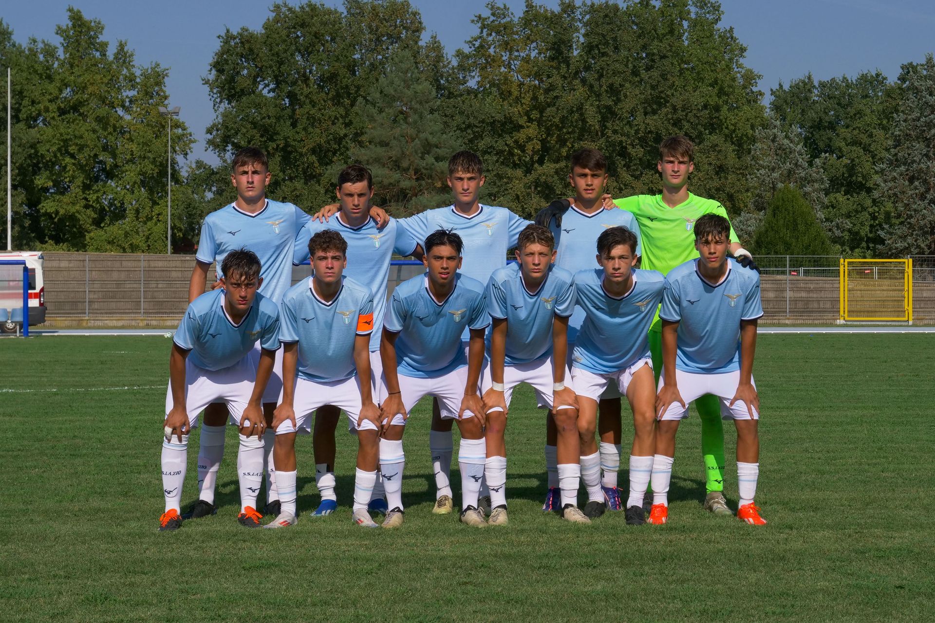 Una squadra di calcio posa su un campo verde, indossando maglie azzurre, pantaloncini bianchi e calzini, sotto un cielo soleggiato.