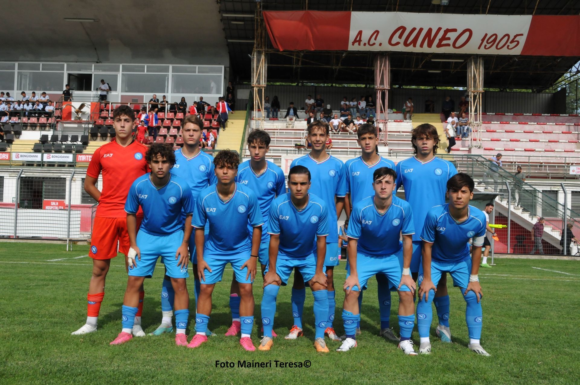 Una squadra di calcio in uniforme blu posa su un campo di fronte a uno stadio con un cartello rosso e bianco.