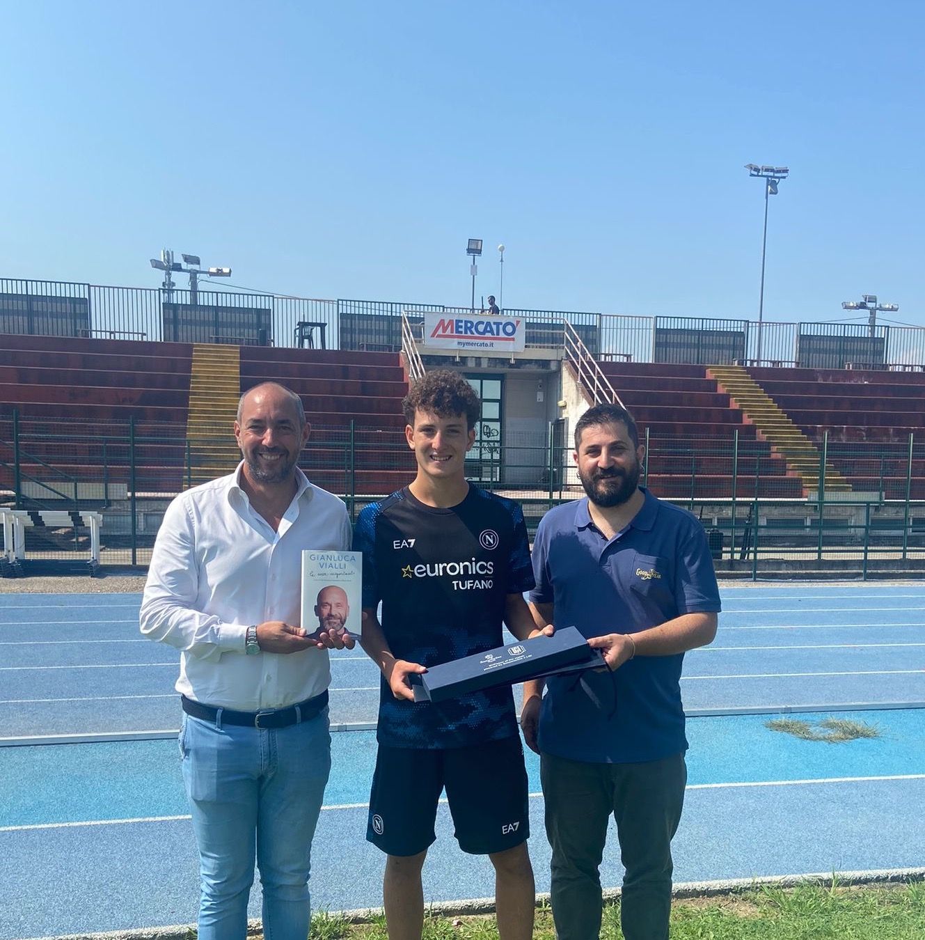 Tre uomini su un campo sportivo: uno tiene in mano un libro, un altro una scatola e la figura centrale sembra essere un giocatore di calcio.