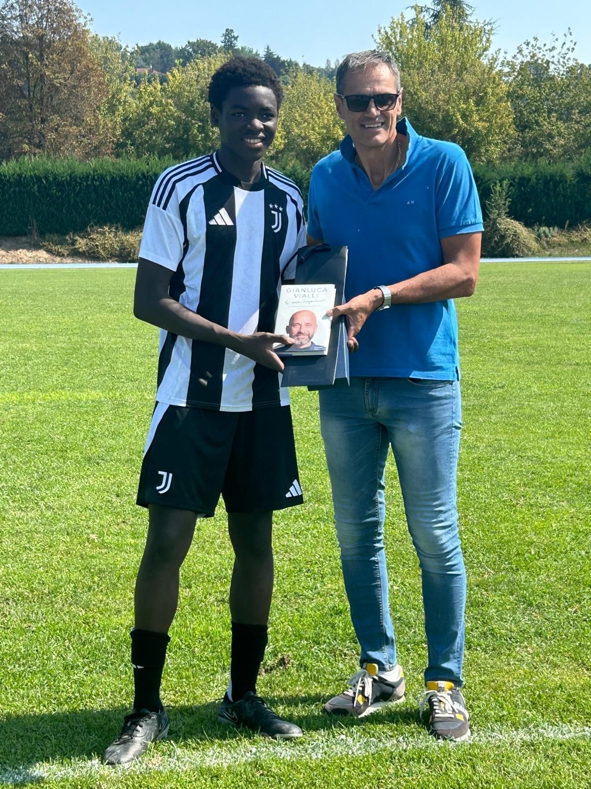 Un calciatore in uniforme bianca e nera tiene in mano un libro, in posa con un uomo in maglietta blu, su un campo erboso.