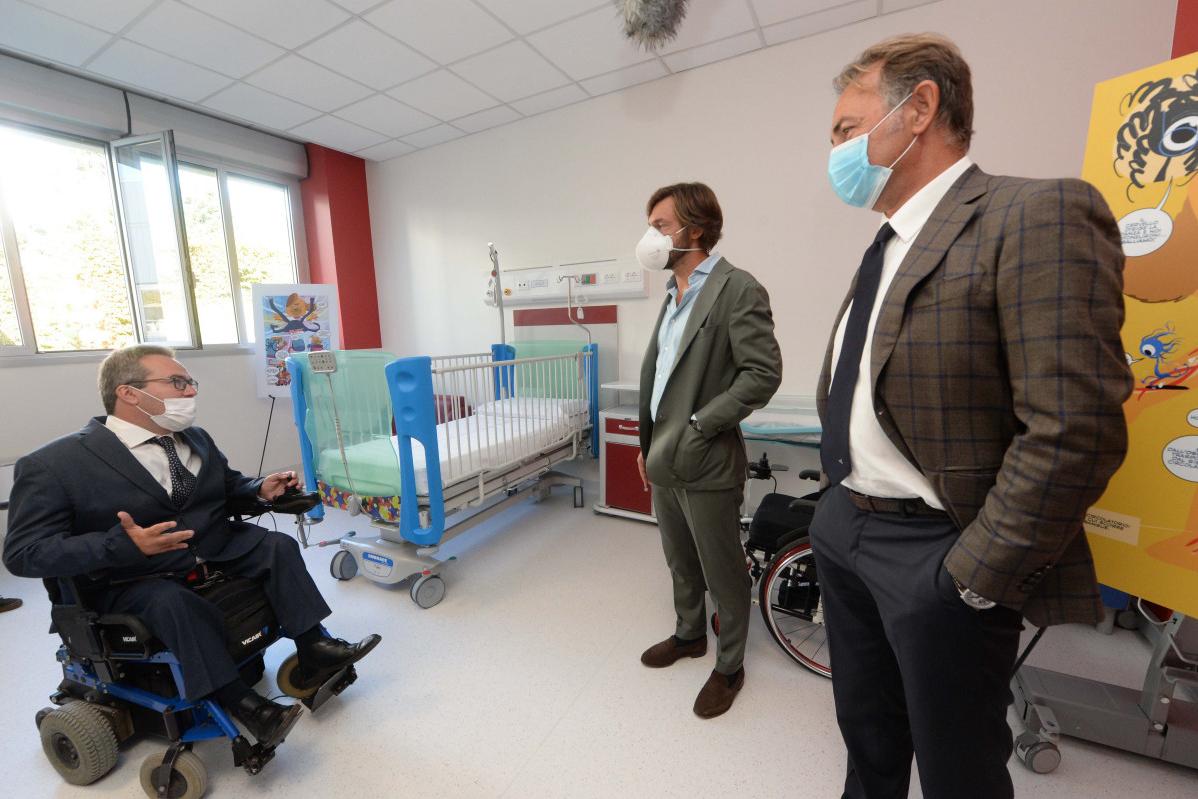 Tre uomini in giacca e cravatta in una stanza d'ospedale, uno su una sedia a rotelle vicino a una culla, tutti con la mascherina.