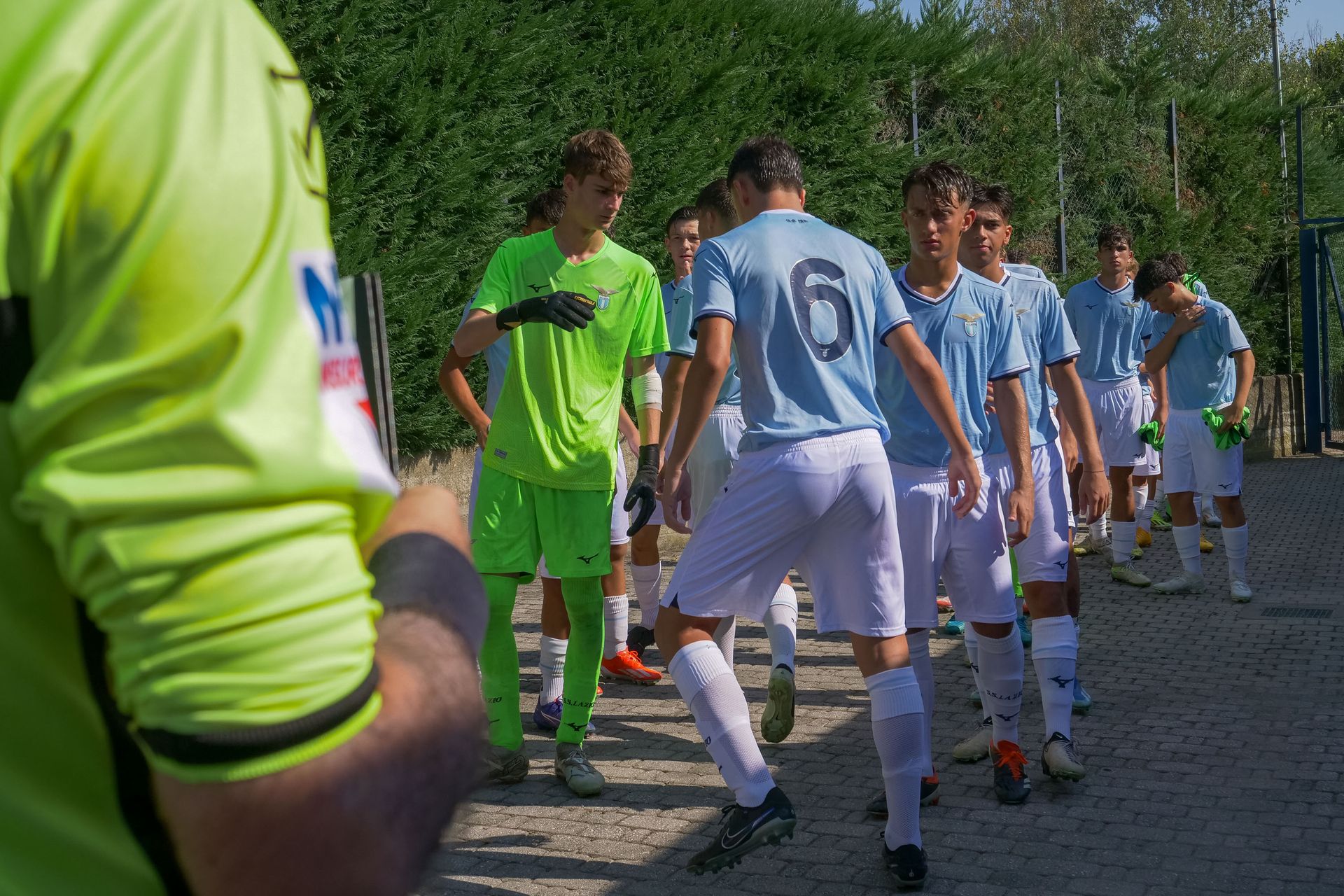 I giocatori di calcio con le maglie azzurre camminano lungo un sentiero verso un campo, di fronte a un allenatore vestito di verde neon.