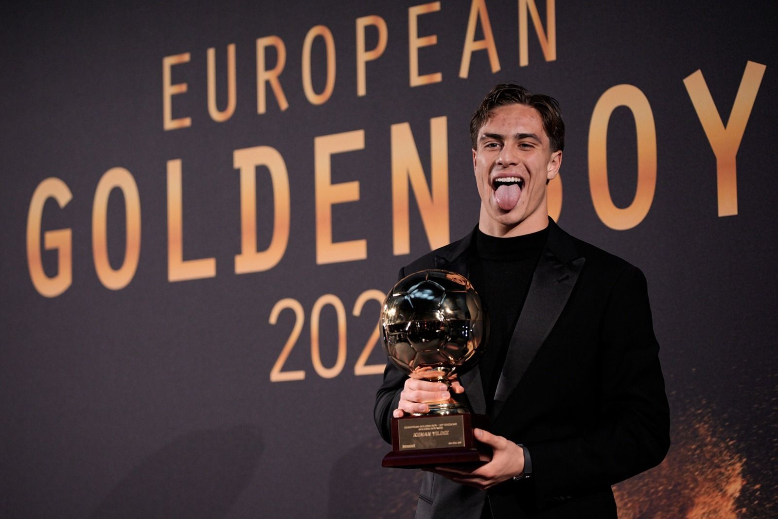 Uomo che tiene in mano il trofeo Golden Boy, tirando fuori la lingua, davanti al cartello