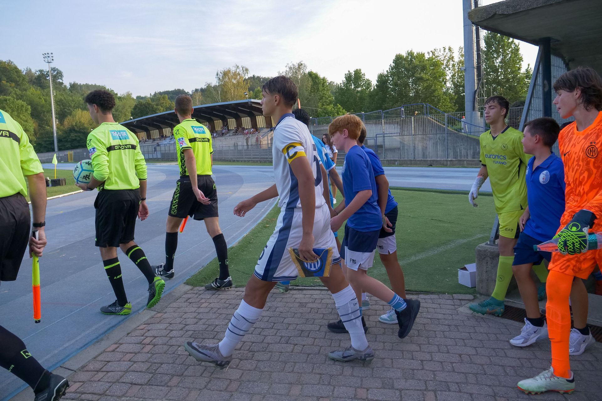 Calciatori e arbitri entrano in campo da un tunnel, sotto un cielo azzurro, indossando le loro uniformi.