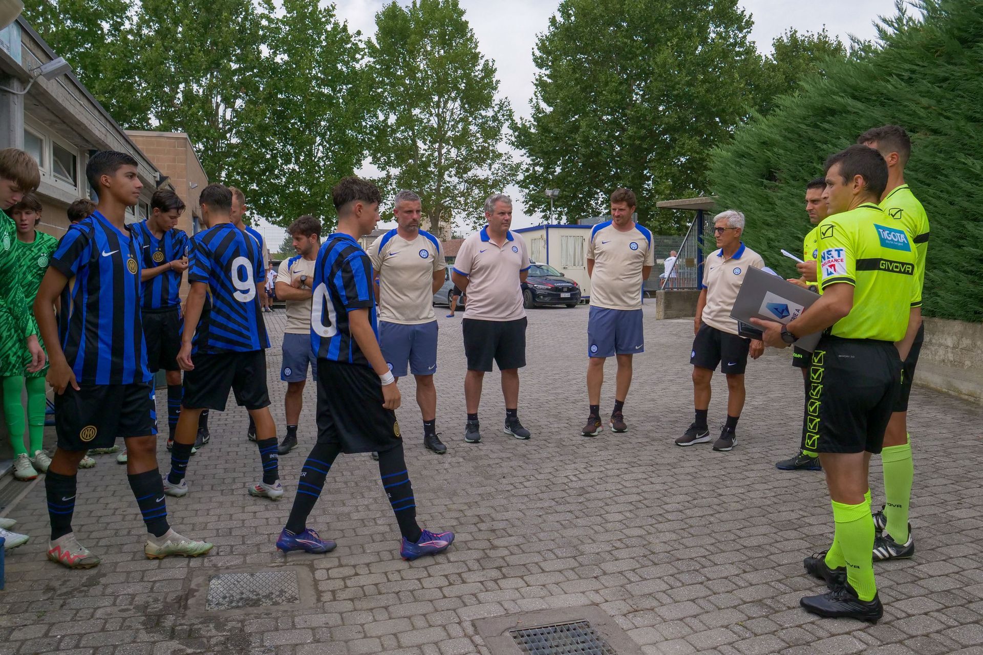 Squadre di calcio in uniforme ascoltano gli arbitri su un'area pavimentata, con alberi ed edifici sullo sfondo.