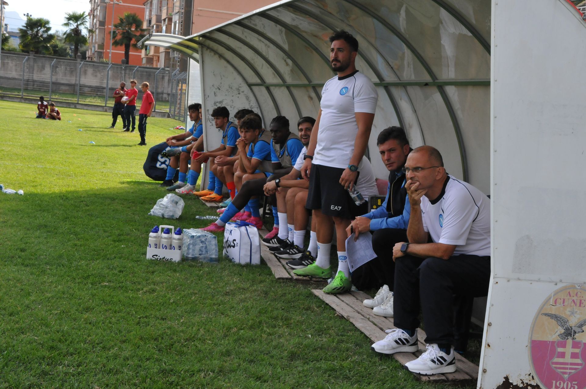 La squadra di calcio è in panchina, sotto una tettoia. L'allenatore è in piedi, gli altri sono seduti, a guardare il campo.