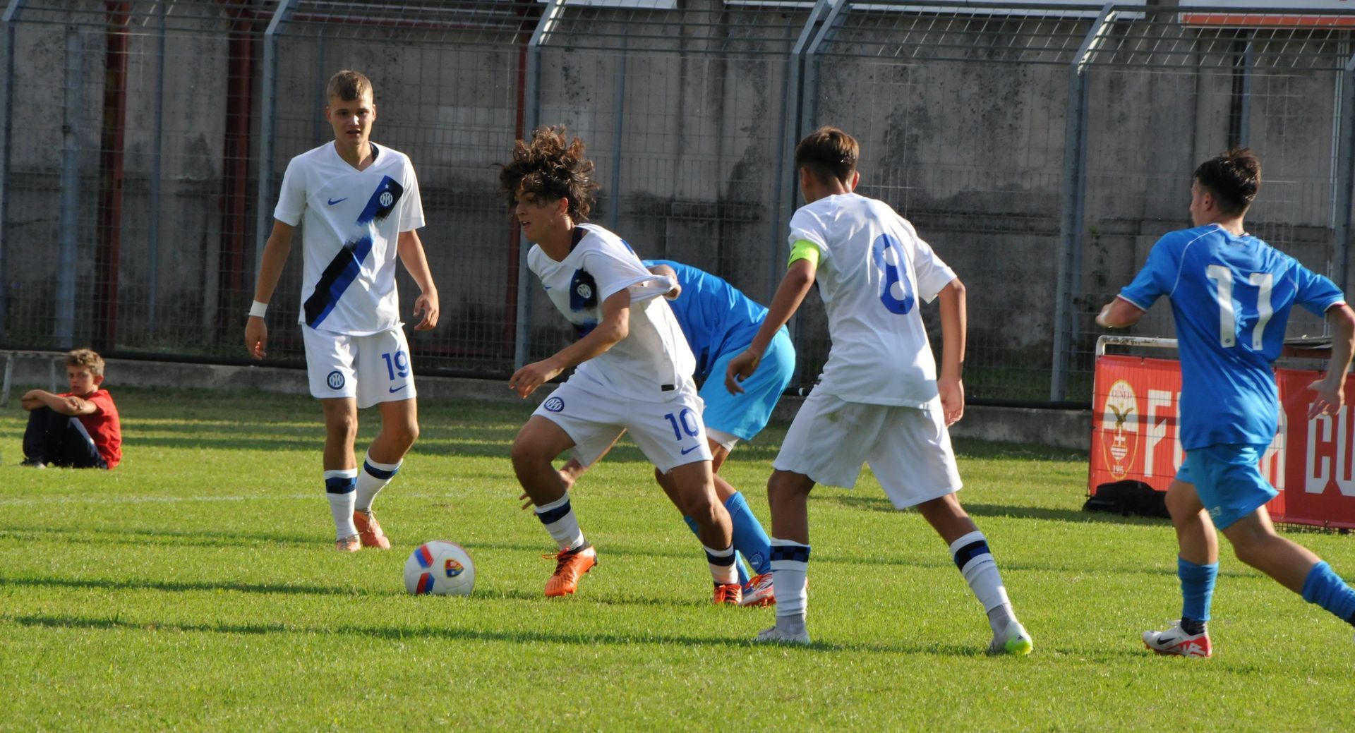Giocatori di calcio con maglie bianche e blu su un campo verde, uno dei quali palleggia.