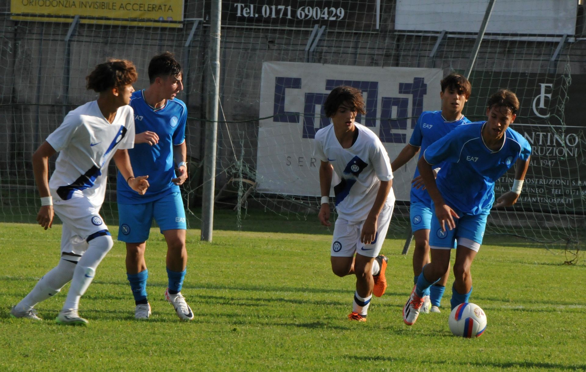 Giocatori di calcio su un campo verde. Giocatori in divisa blu e bianca, uno dei quali calcia un pallone.