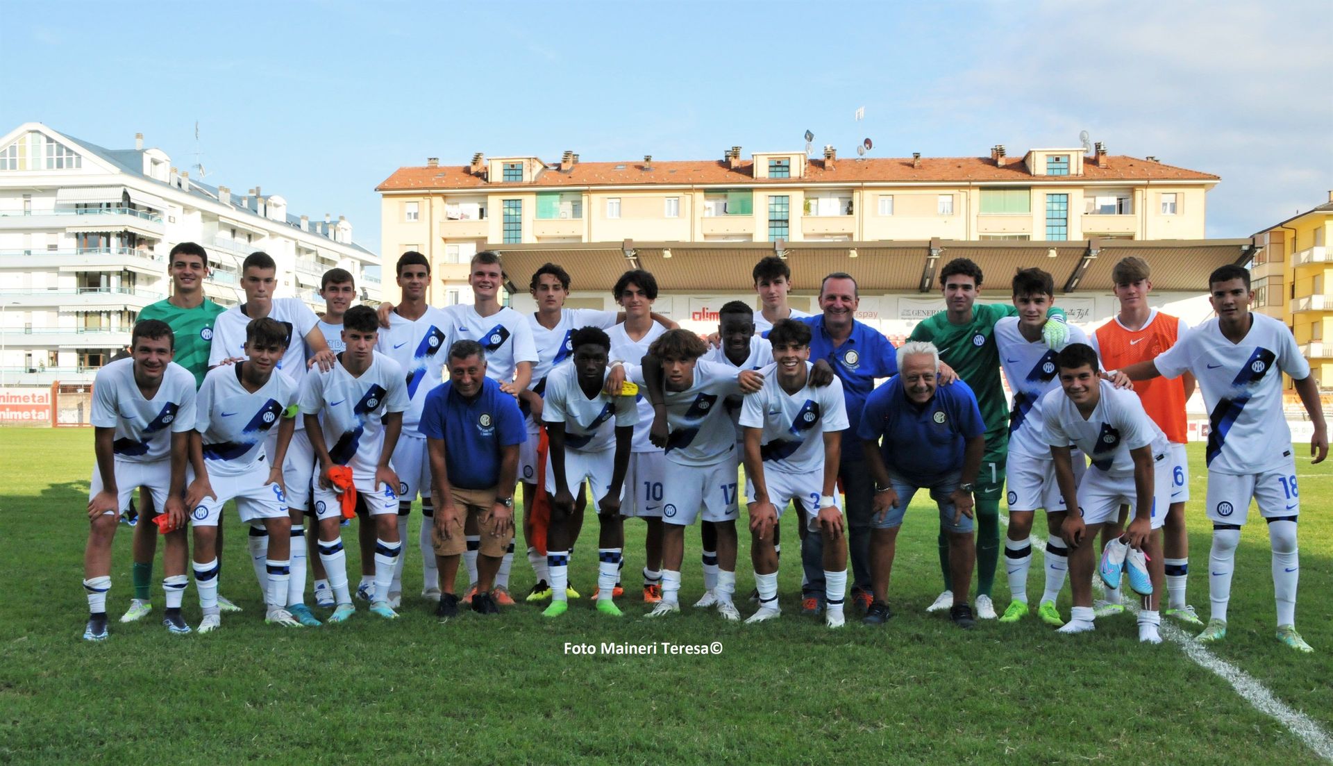 La squadra di calcio posa su un campo soleggiato. I giocatori indossano divise bianche e blu, gli allenatori posano con loro davanti agli edifici.