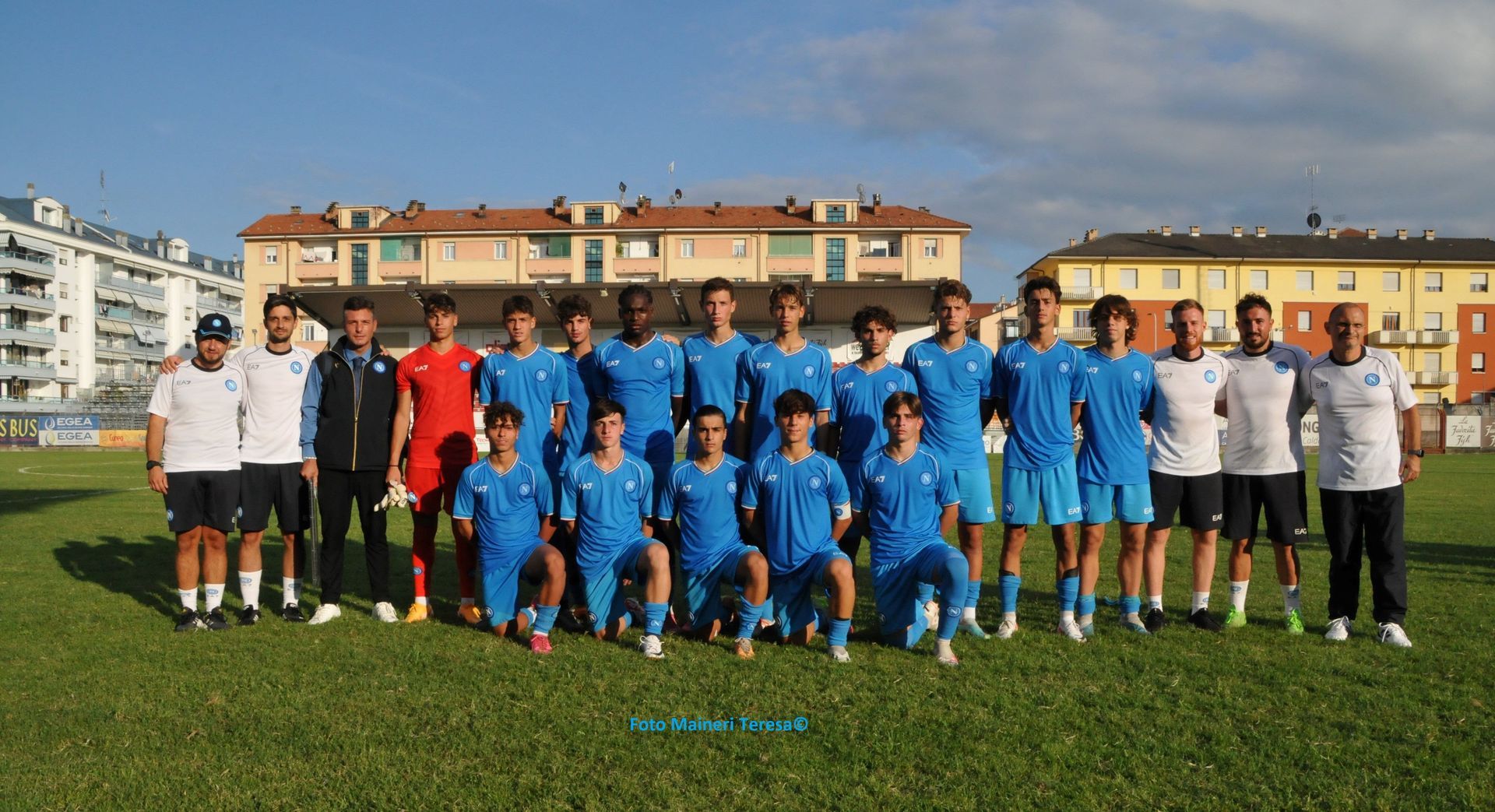 Una squadra di calcio in uniforme blu posa su un campo verde di fronte agli edifici in una giornata di sole.