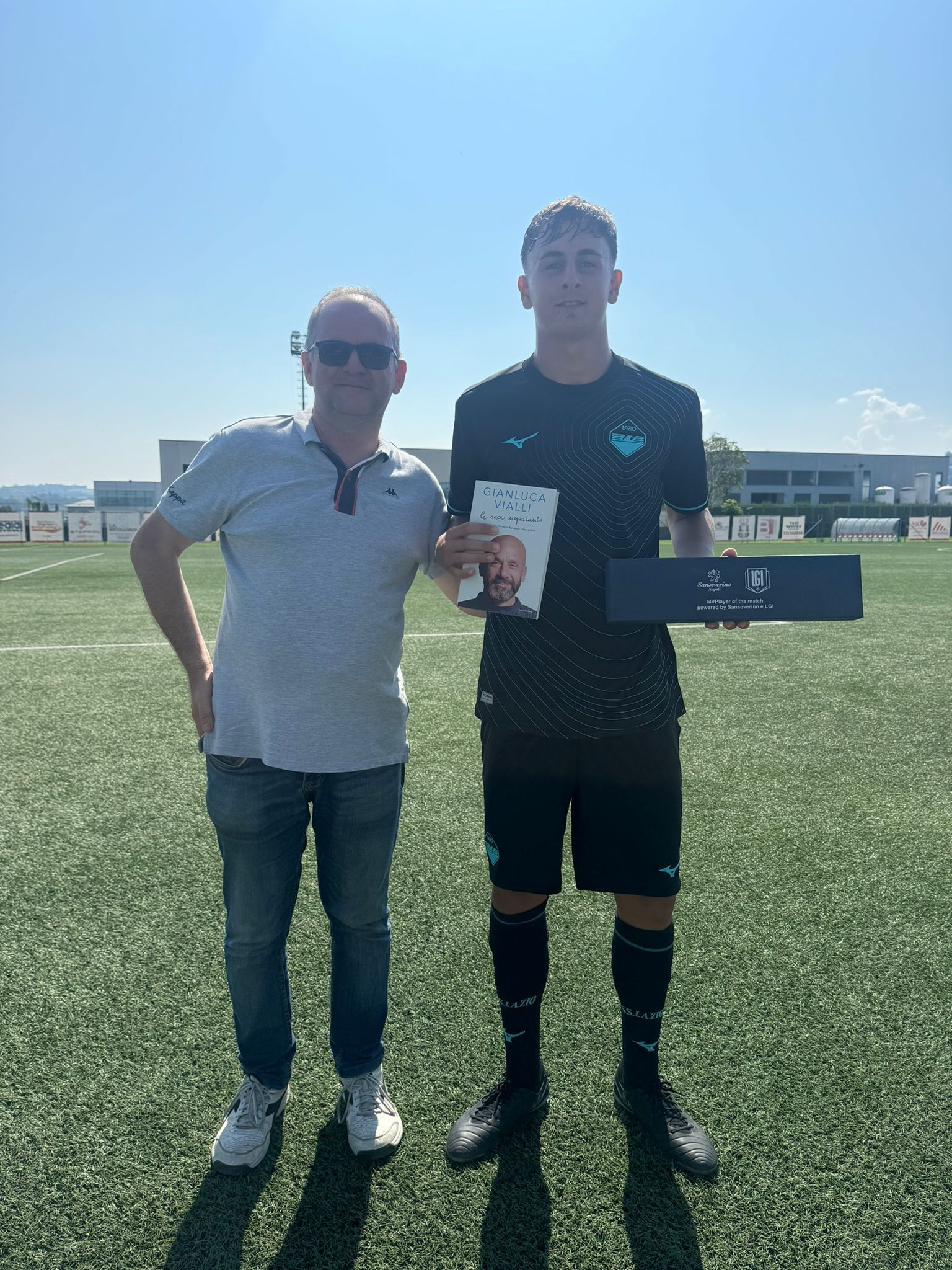 Un uomo consegna un premio a un giocatore di calcio su un campo verde sotto un cielo soleggiato.