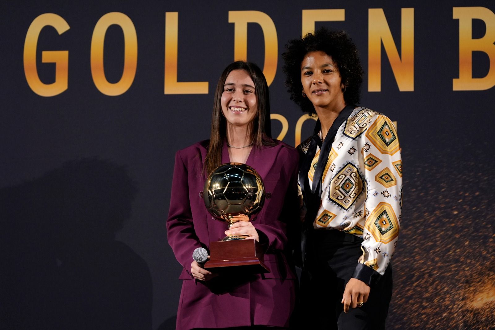 Due donne sul palco, una delle quali regge un trofeo dorato. Dietro di loro c'è uno sfondo dorato con la scritta