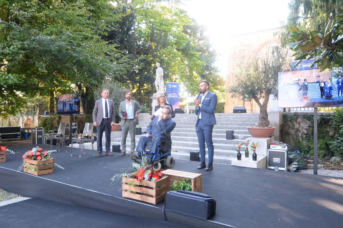 Un gruppo di uomini su un palco all'aperto. Uno è su una sedia a rotelle, gli altri sono in piedi. Le decorazioni includono piante e schermi.