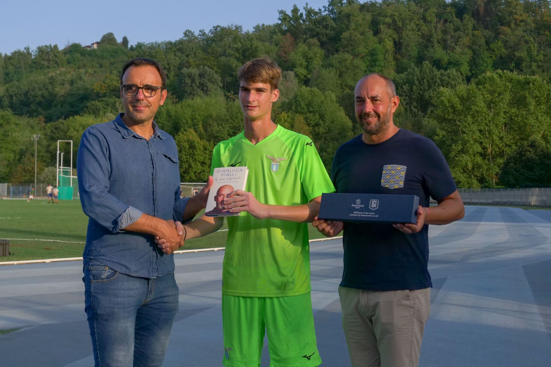 Tre persone su una pista. Un giovane in uniforme da calcio verde lime riceve un premio da due uomini.