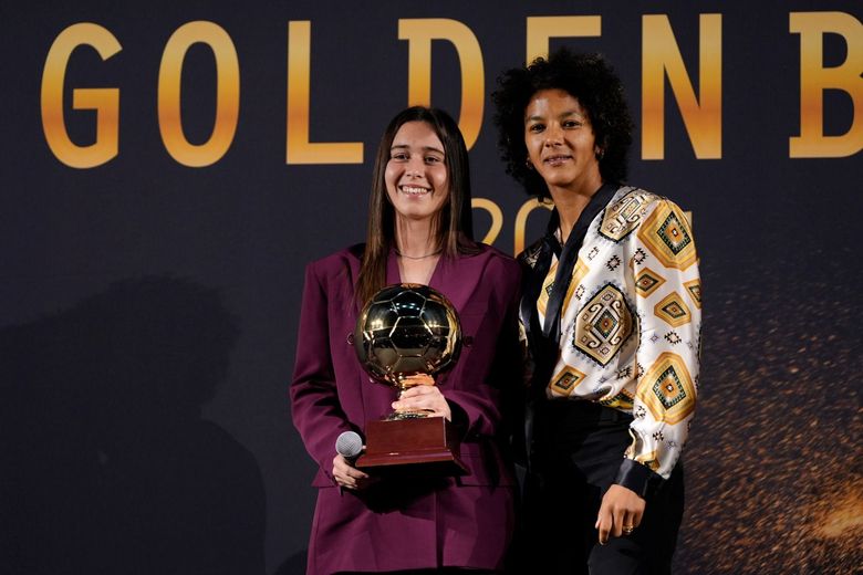 Due donne sul palco, una delle quali regge un trofeo dorato. Dietro di loro c'è uno sfondo dorato con la scritta 