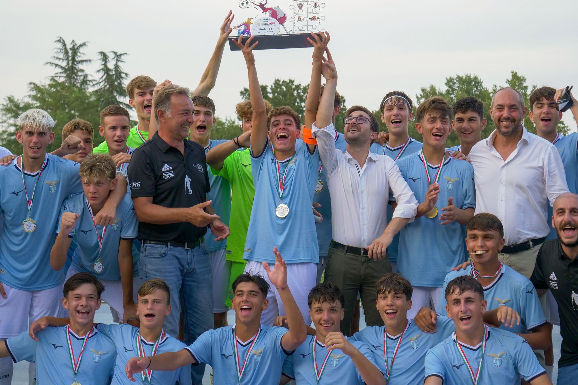Squadra di calcio che festeggia una vittoria, con in mano un trofeo, divise azzurre, ambientazione all'aperto.
