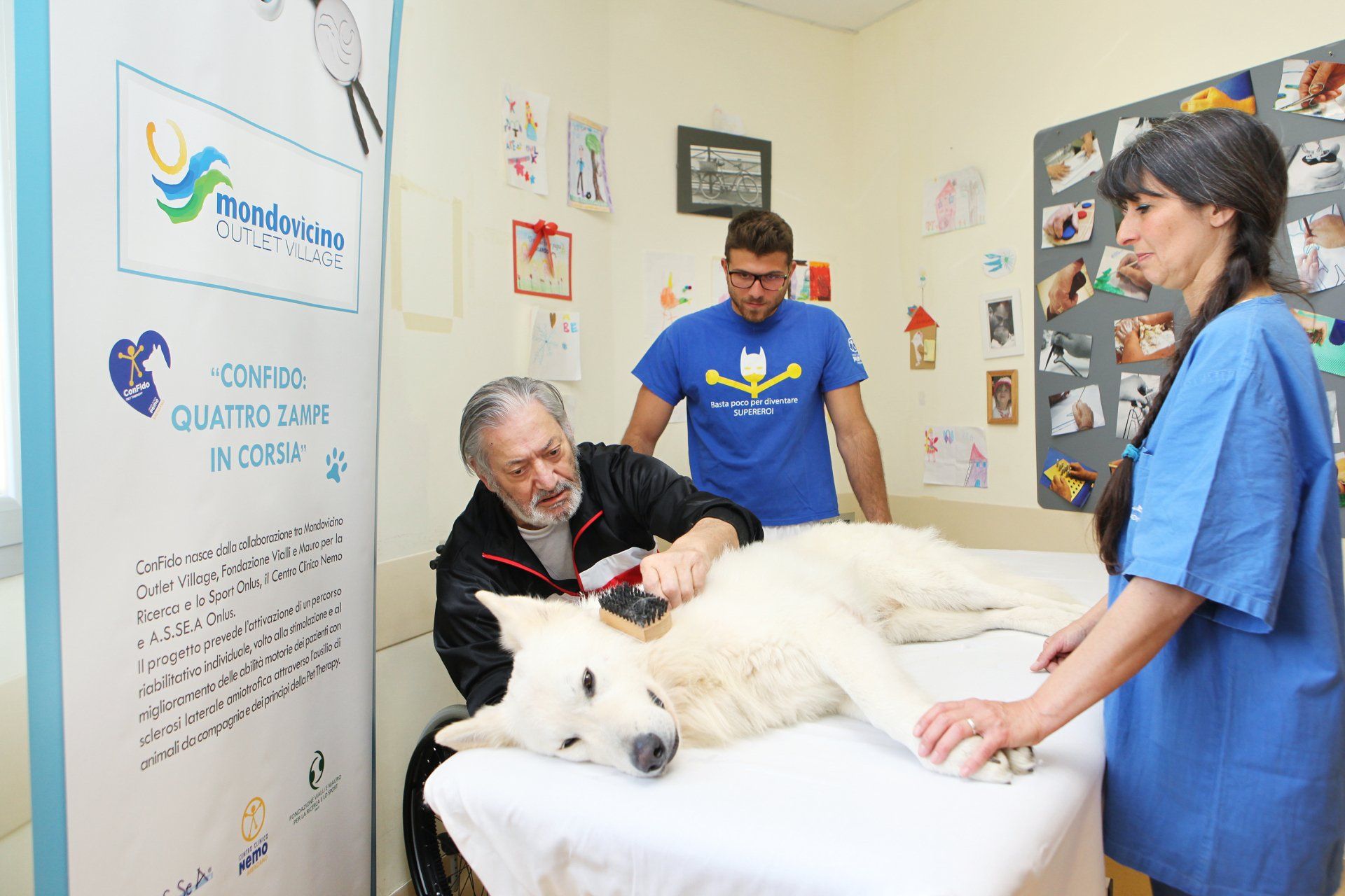 Un uomo sfiora un cane sul lettino da visita; altri due osservano in clinica con un'opera d'arte.