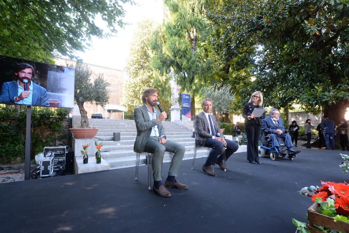 Evento con relatori all'aperto; un uomo parla, altri sono seduti, sullo schermo è mostrato un altro relatore.