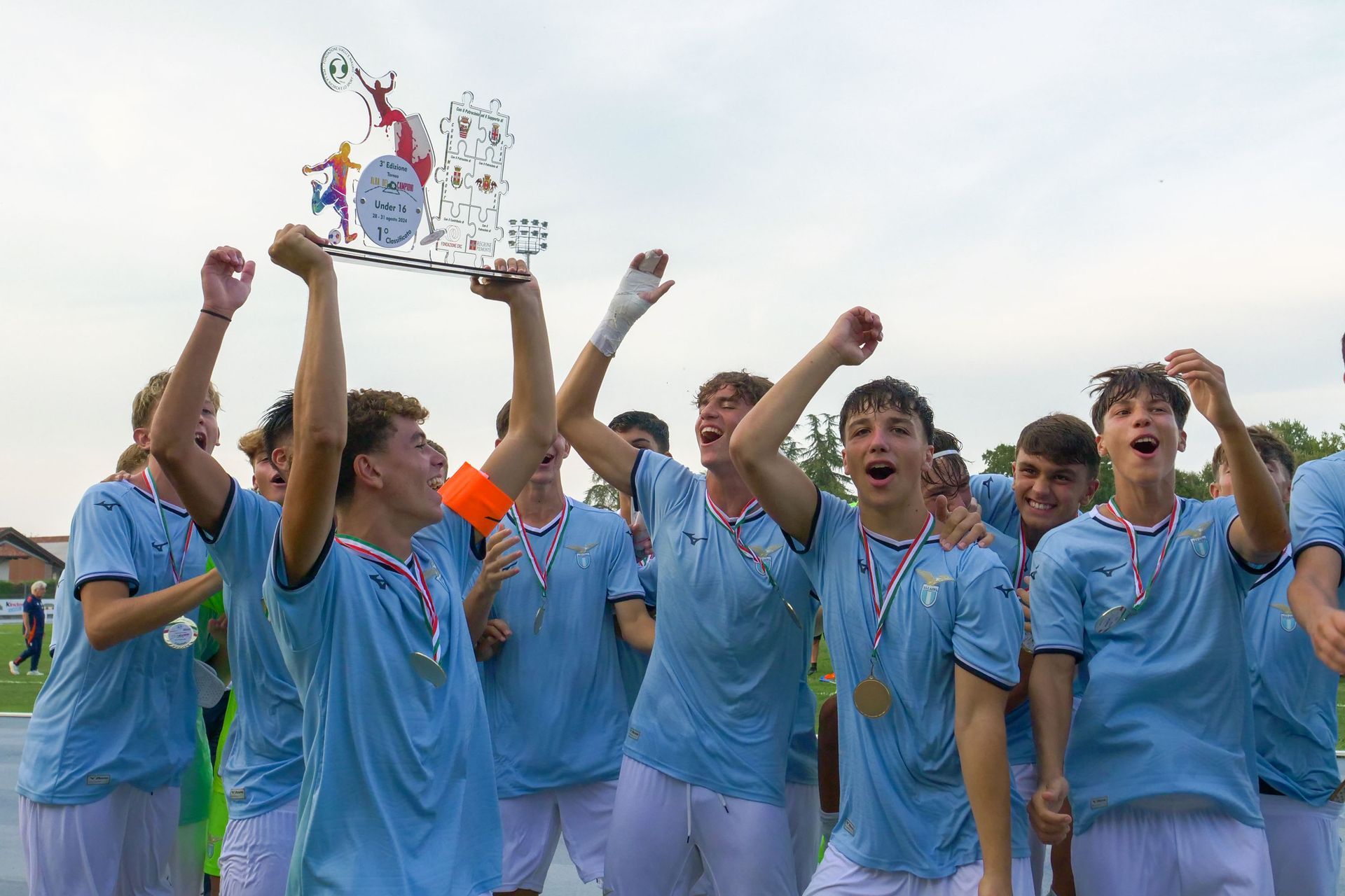 La squadra di calcio festeggia una vittoria, tenendo in mano un trofeo e delle medaglie. Maglie azzurre, espressioni di giubilo, cornice all'aperto.