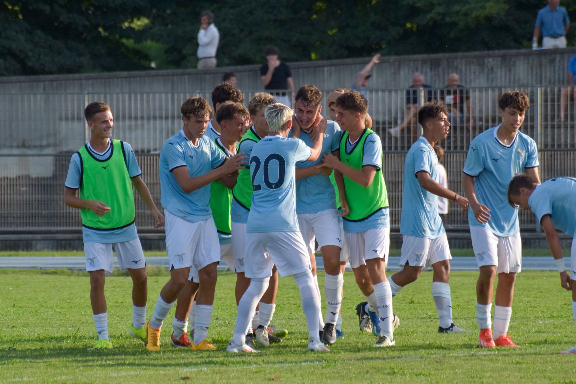 La squadra di calcio con le maglie azzurre festeggia un gol su un campo verde. I giocatori si abbracciano, esultando con entusiasmo.
