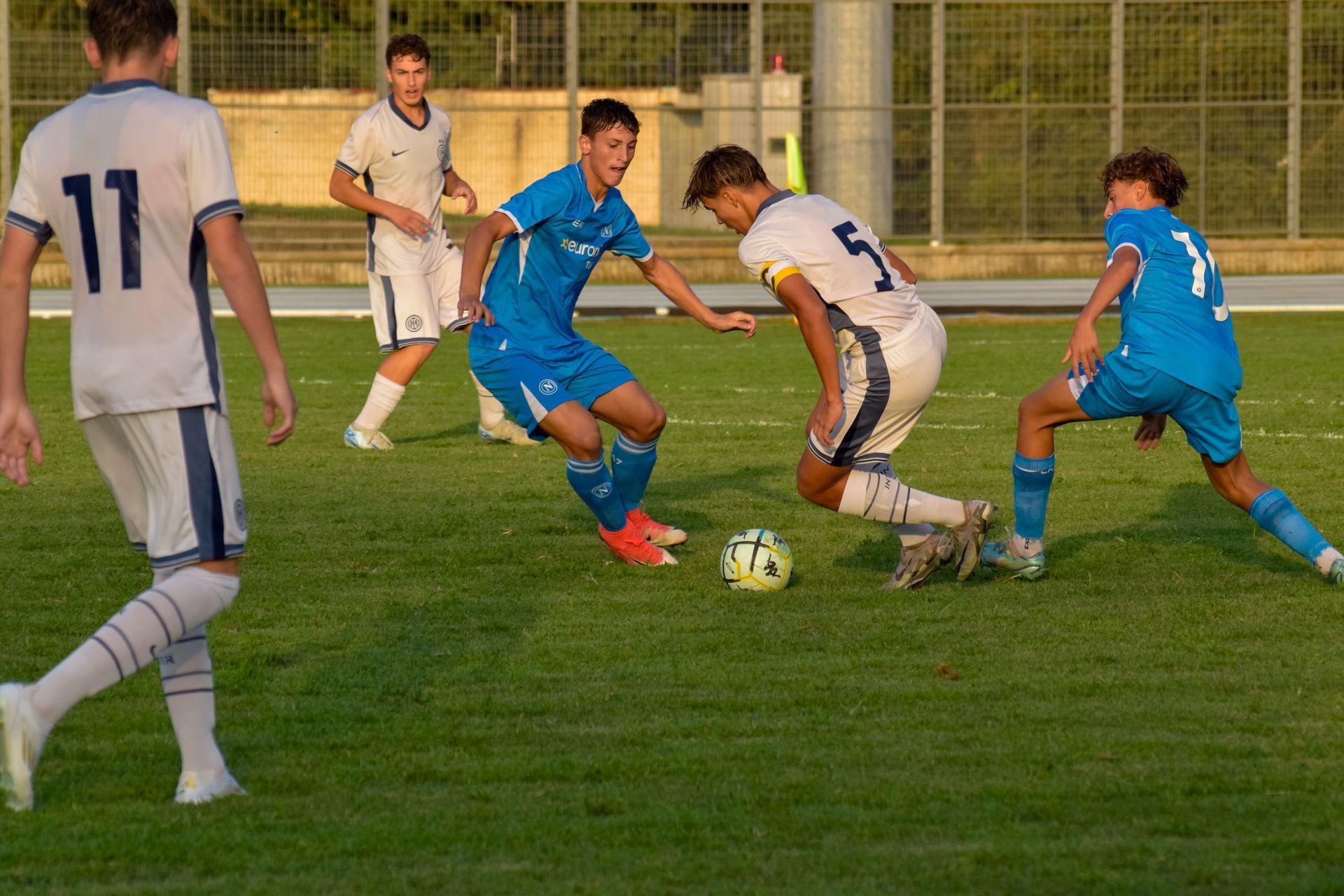Giocatori di calcio su un campo verde; un giocatore in blu calcia la palla, altri con maglie blu e bianche nelle vicinanze.