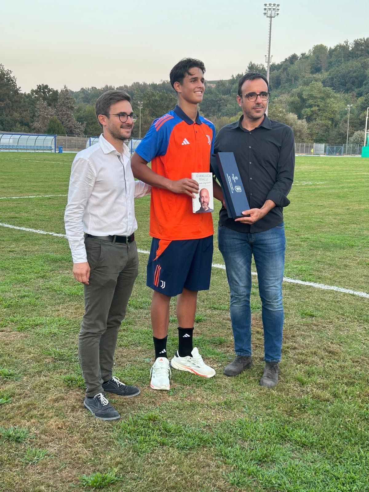Tre persone su un campo verde. Una persona con una maglia arancione/blu tiene in mano un libro e un regalo. Altre due persone la affiancano.