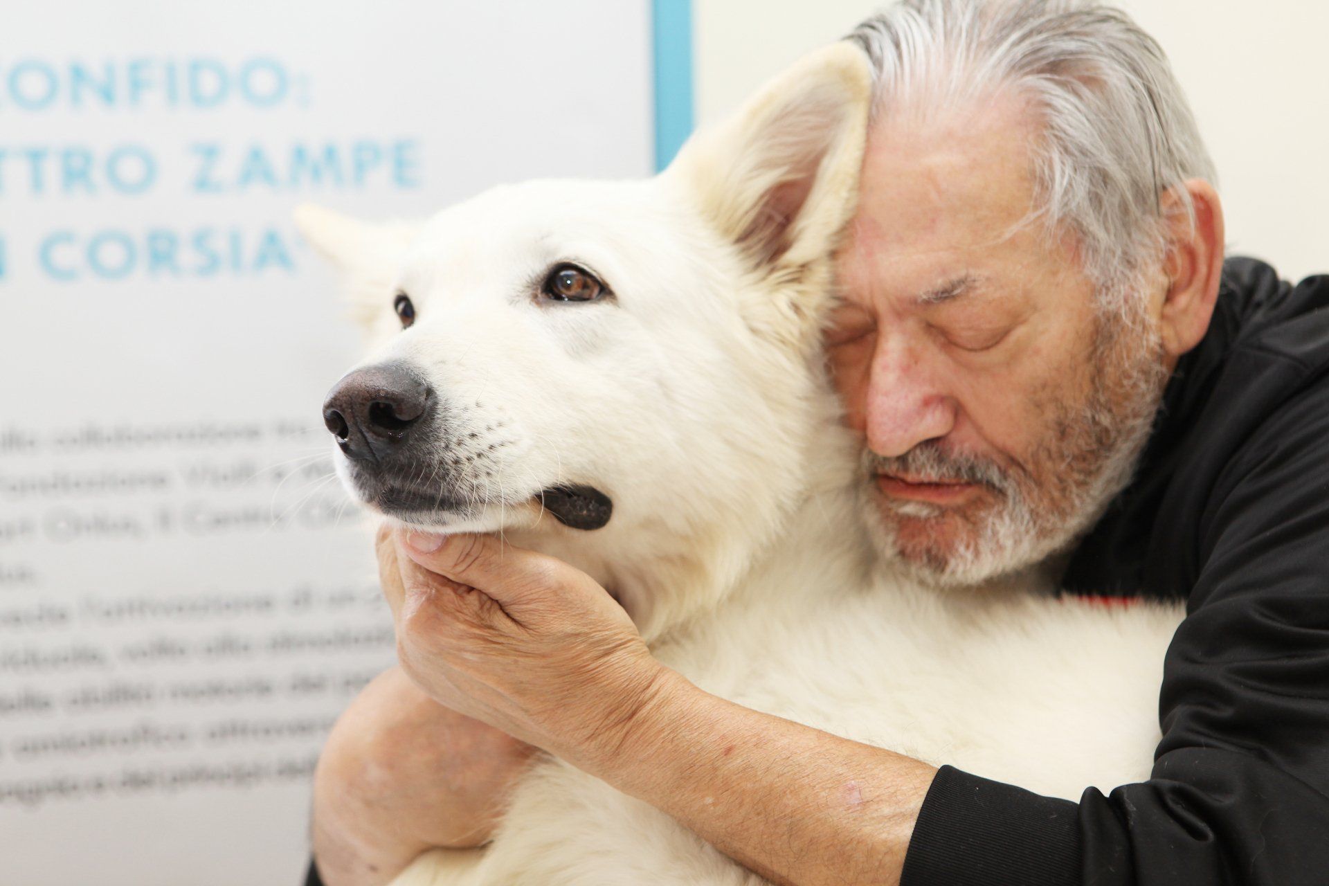Un uomo abbraccia un cane bianco, entrambi con gli occhi chiusi, vicino a un cartello.