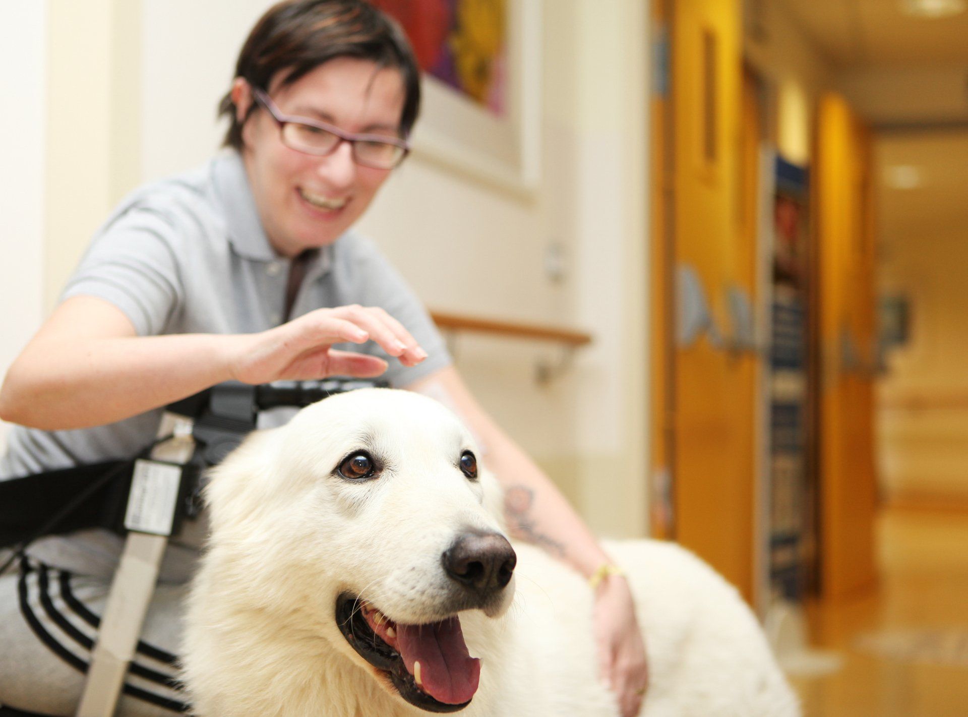 Una donna con gli occhiali accarezza un cane bianco in un corridoio. Entrambi sorridono.
