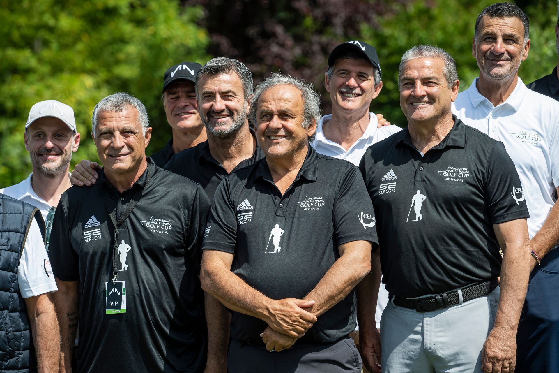 Gruppo di uomini in posa per una foto all'aperto, davanti ad alberi verdi. Alcuni indossano camicie nere e sorridono.