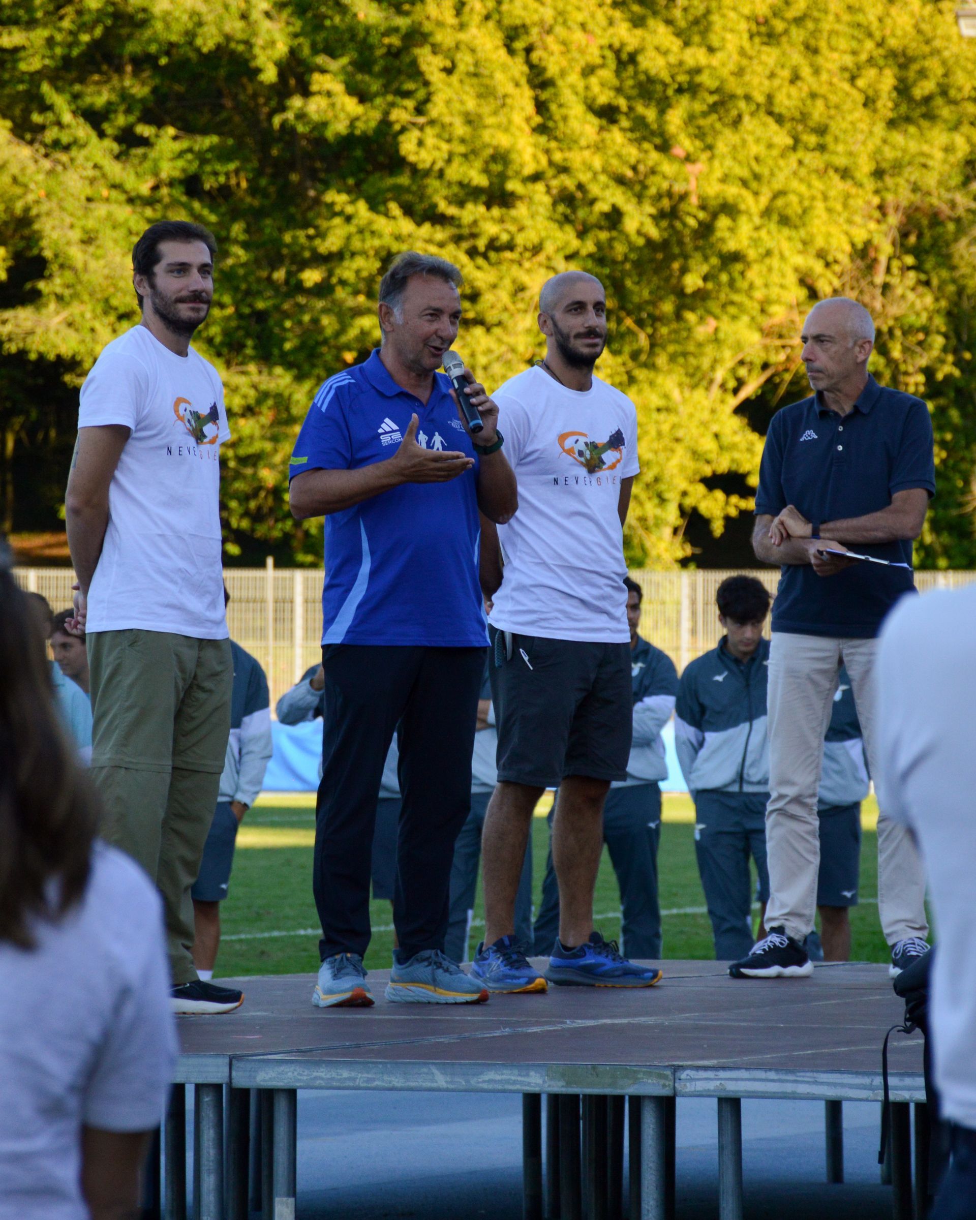 Quattro uomini sono in piedi su un palco, uno dei quali parla al microfono. Sono all'aperto, con un campo da calcio e alberi sullo sfondo.