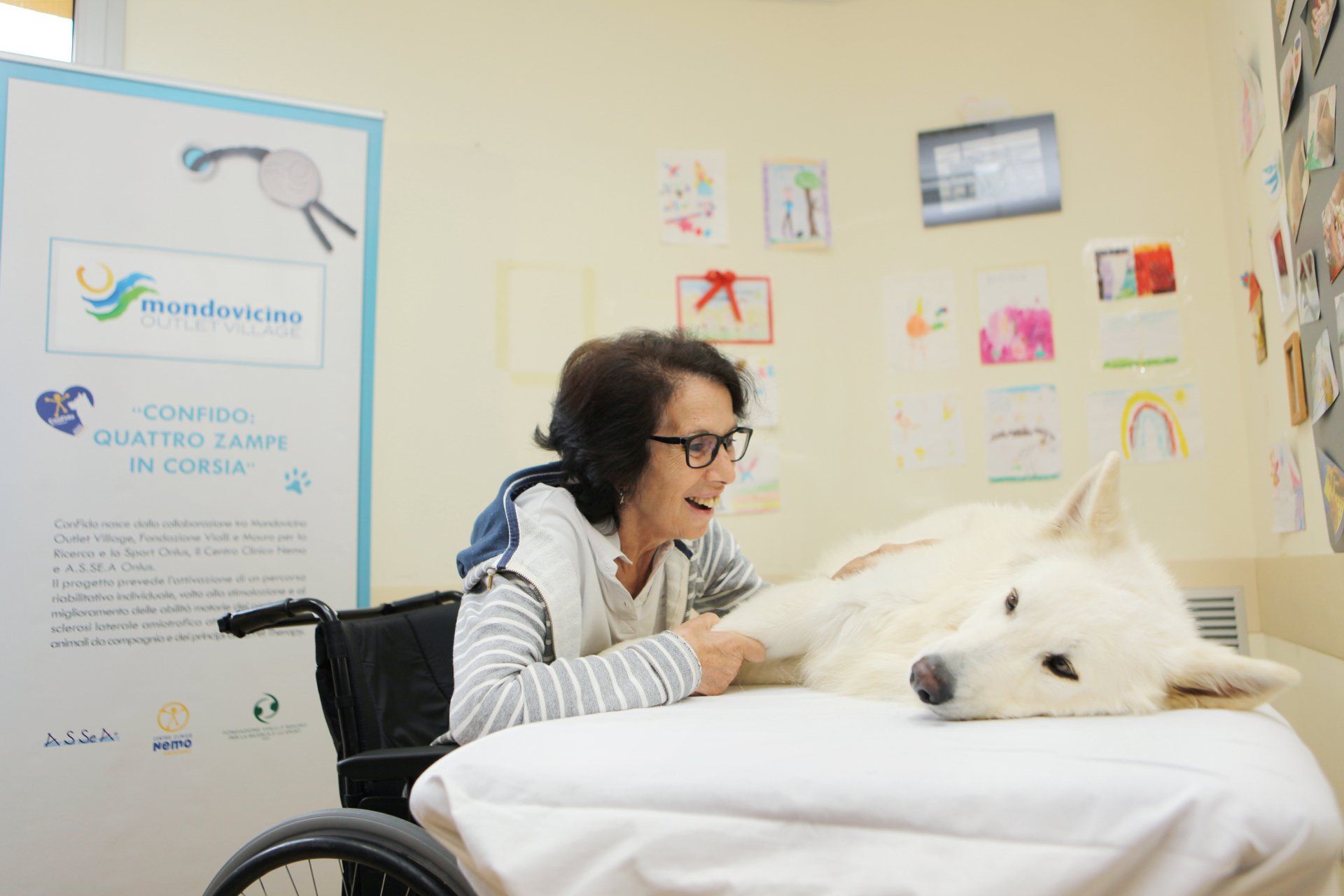 Donna in sedia a rotelle che accarezza un grosso cane bianco sdraiato su un tavolo. Stanza con disegni e un cartello.