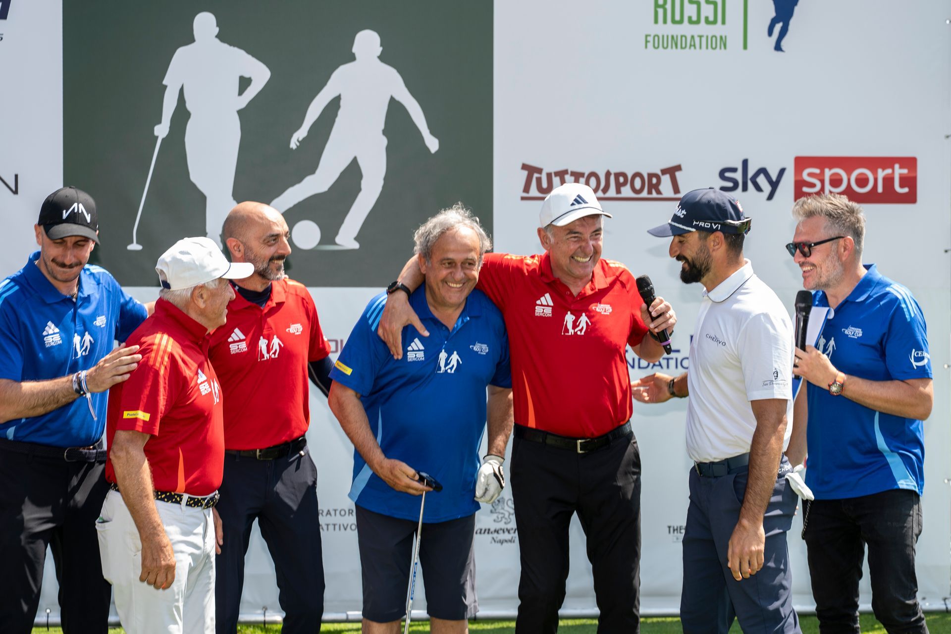 Un gruppo di uomini sorridenti e chiacchieranti su un campo da golf, alcuni abbracciati e con in mano un microfono.