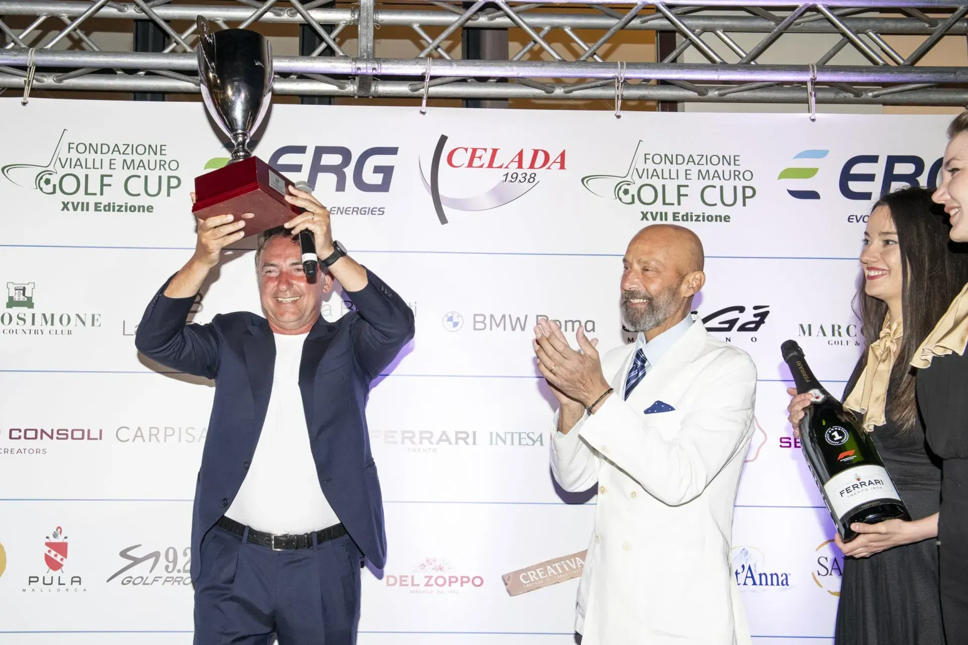 Uomo che tiene in mano un trofeo, festeggia con altri durante una cerimonia di premiazione, con i loghi degli sponsor visibili.