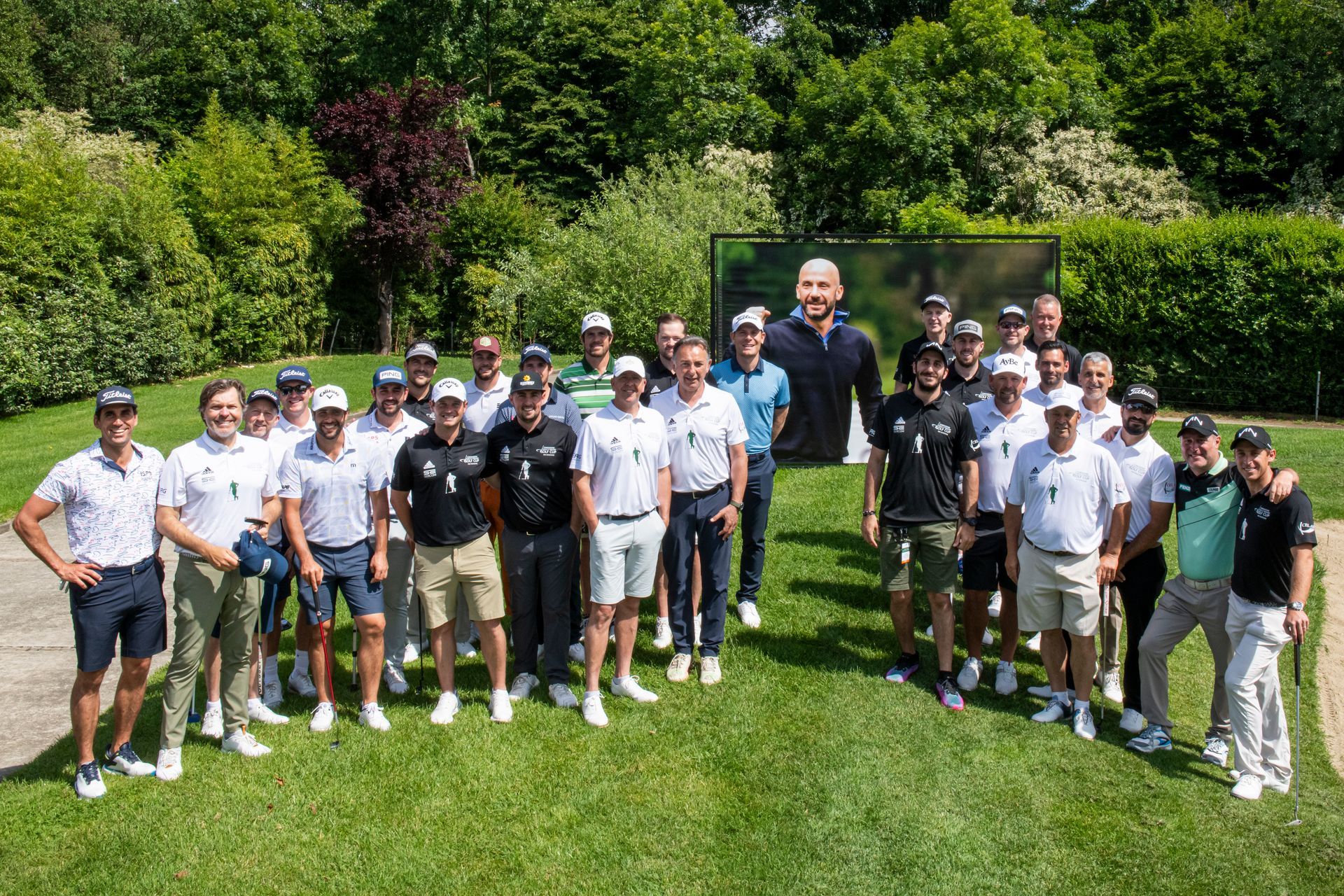 Un gruppo di uomini in tenuta da golf posano sorridenti su un green. Sullo sfondo, una grande foto di un uomo. Giornata di sole.