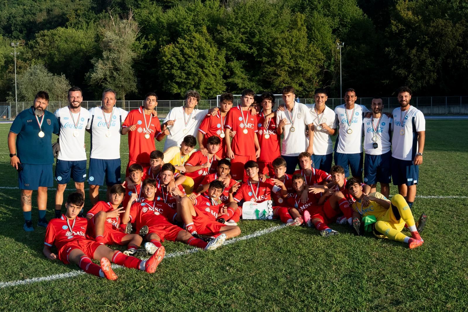 Quattro uomini sono in piedi su un palco, uno dei quali parla al microfono. Sono all'aperto, con un campo da calcio e alberi sullo sfondo.