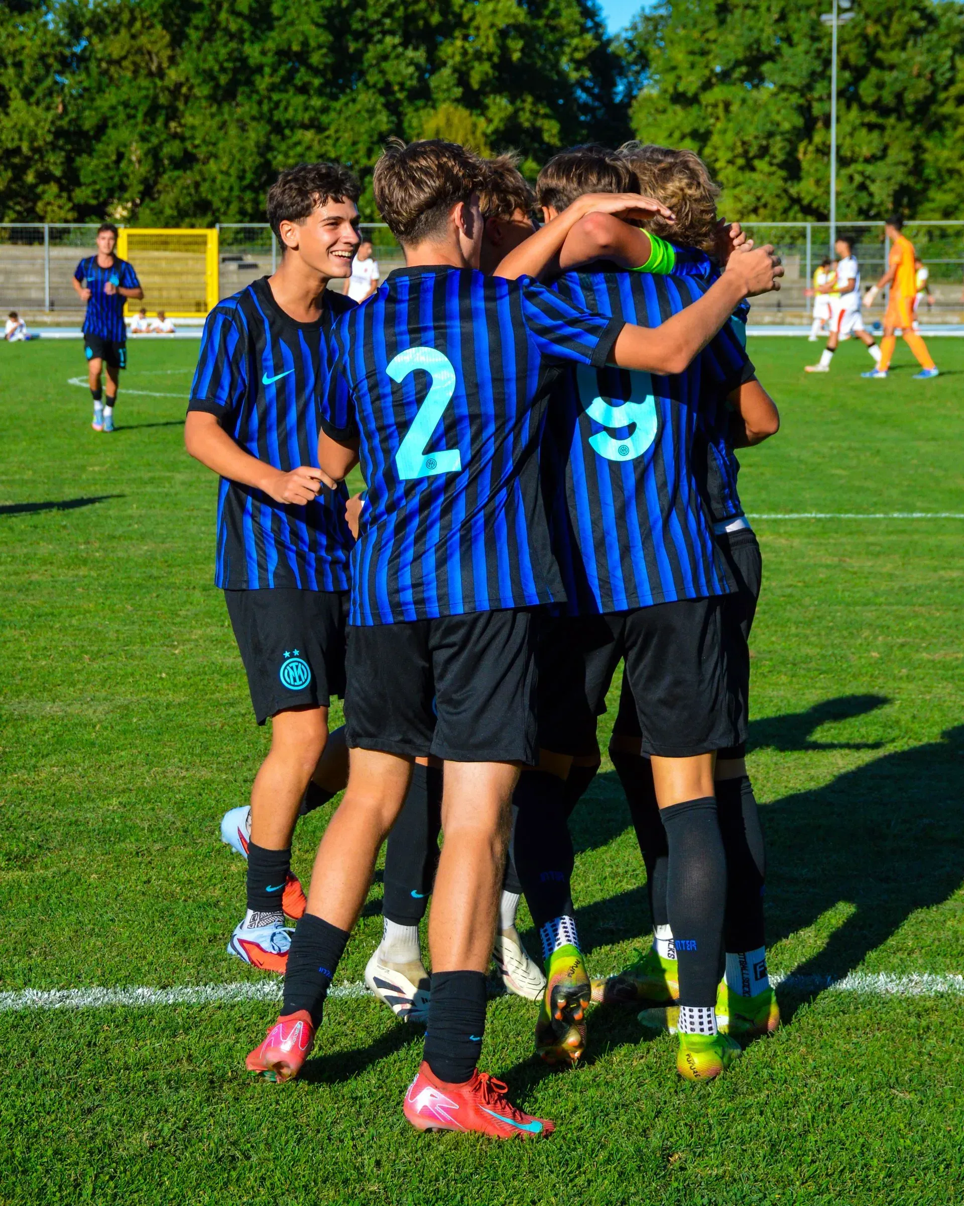 La squadra di calcio si riunisce, festeggiando un gol su un campo verde. Giocatori in divisa blu e nera, uno sorride. Soleggiato scenario all'aperto.