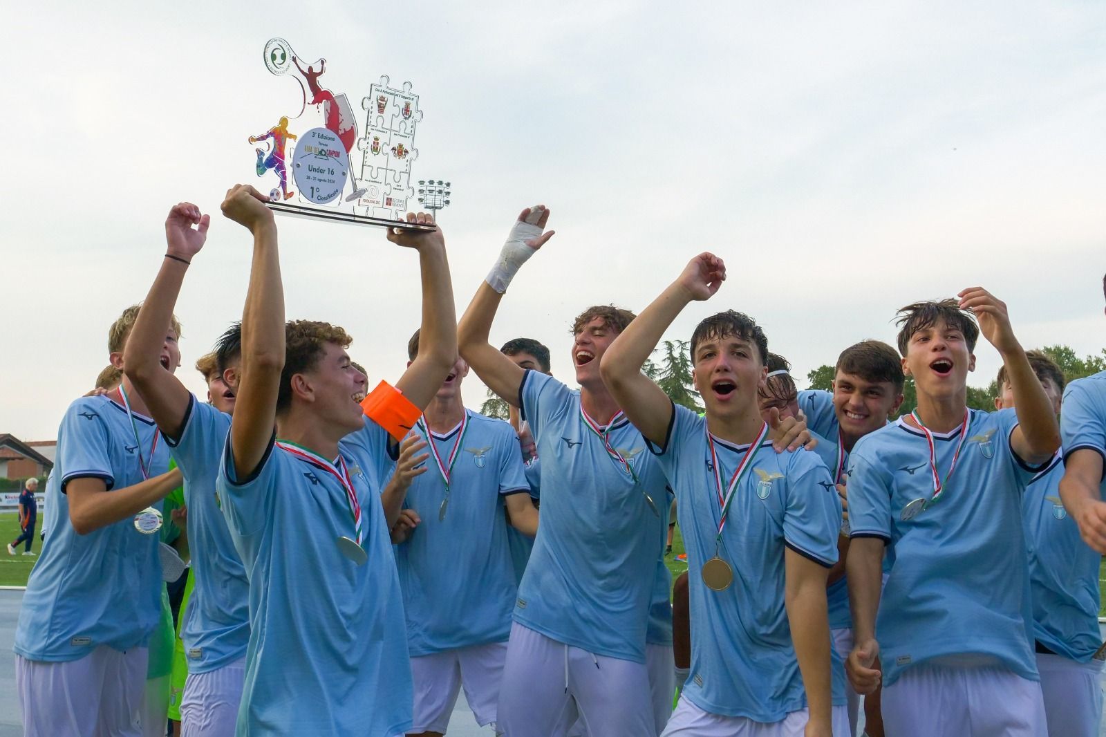 Squadra di calcio che festeggia una vittoria, con in mano un trofeo e delle medaglie. Divise azzurre, all'aperto.