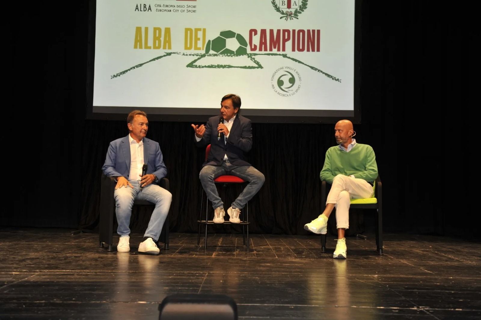 Tre uomini sono seduti su un palco e discutono su uno schermo su cui è scritto
