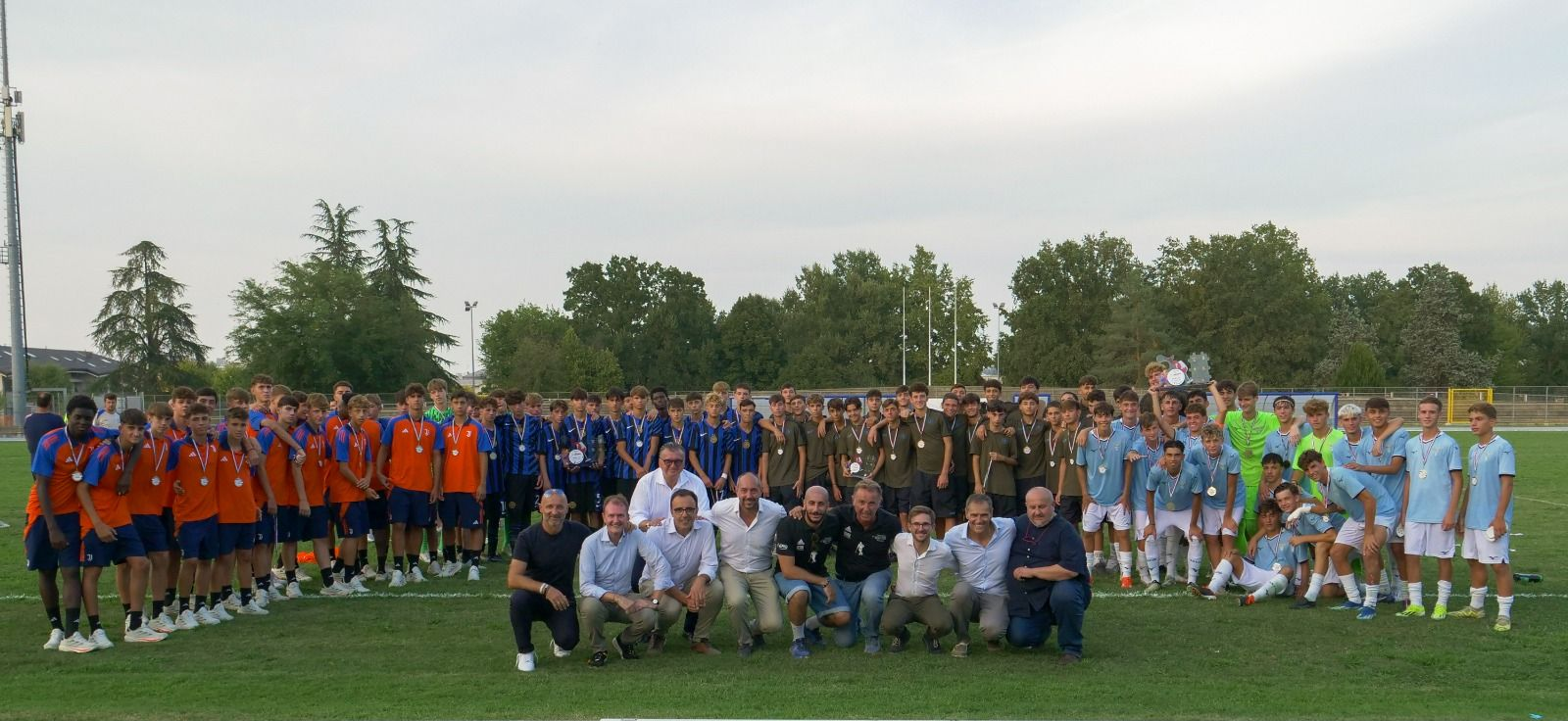 Squadre di calcio con maglie arancioni e blu affiancano un gruppo di persone, alcune in uniforme militare, su un campo verde.