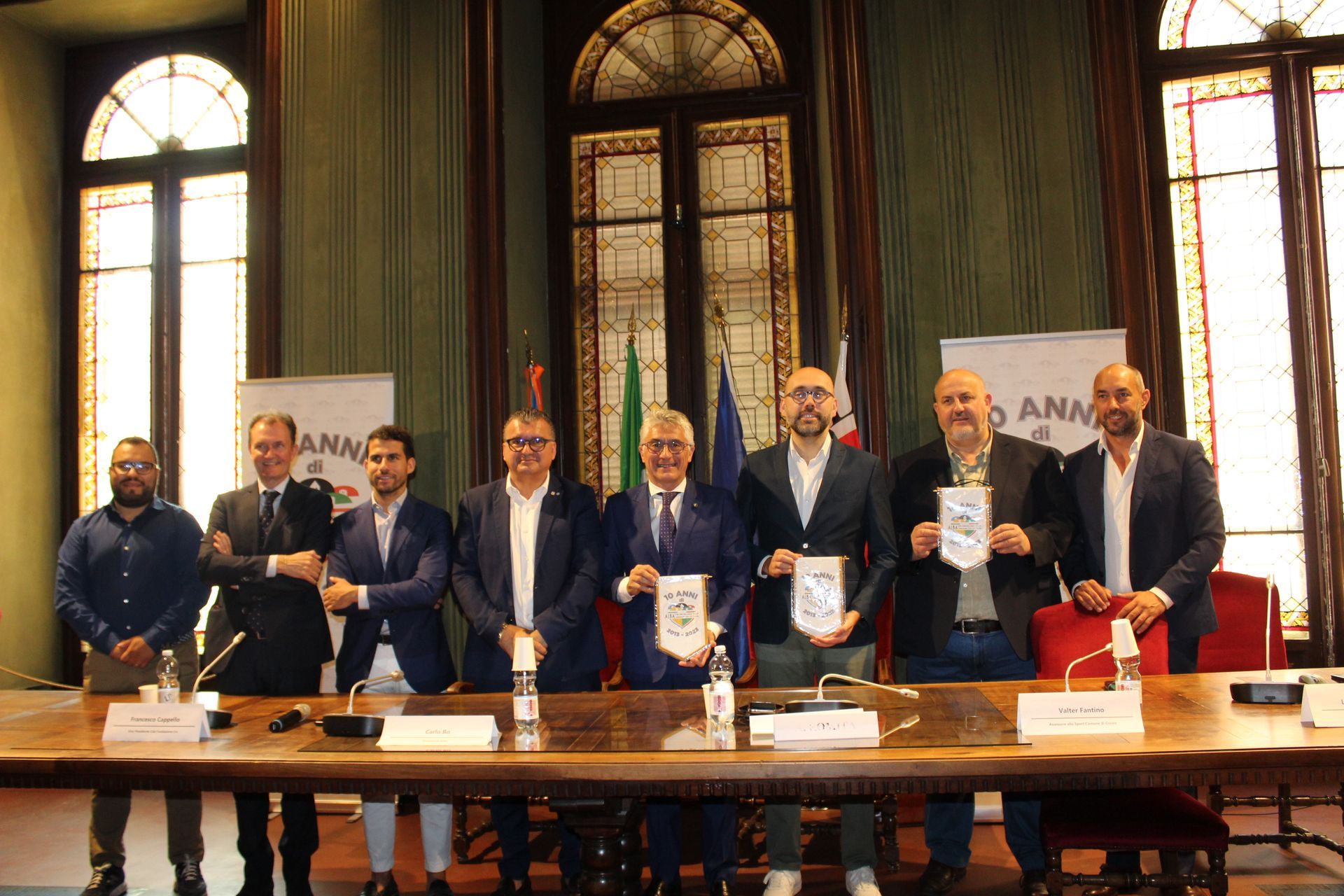 Un gruppo di uomini con dei documenti in mano seduti a un tavolo. Si trovano in una stanza con grandi finestre e bandiere.