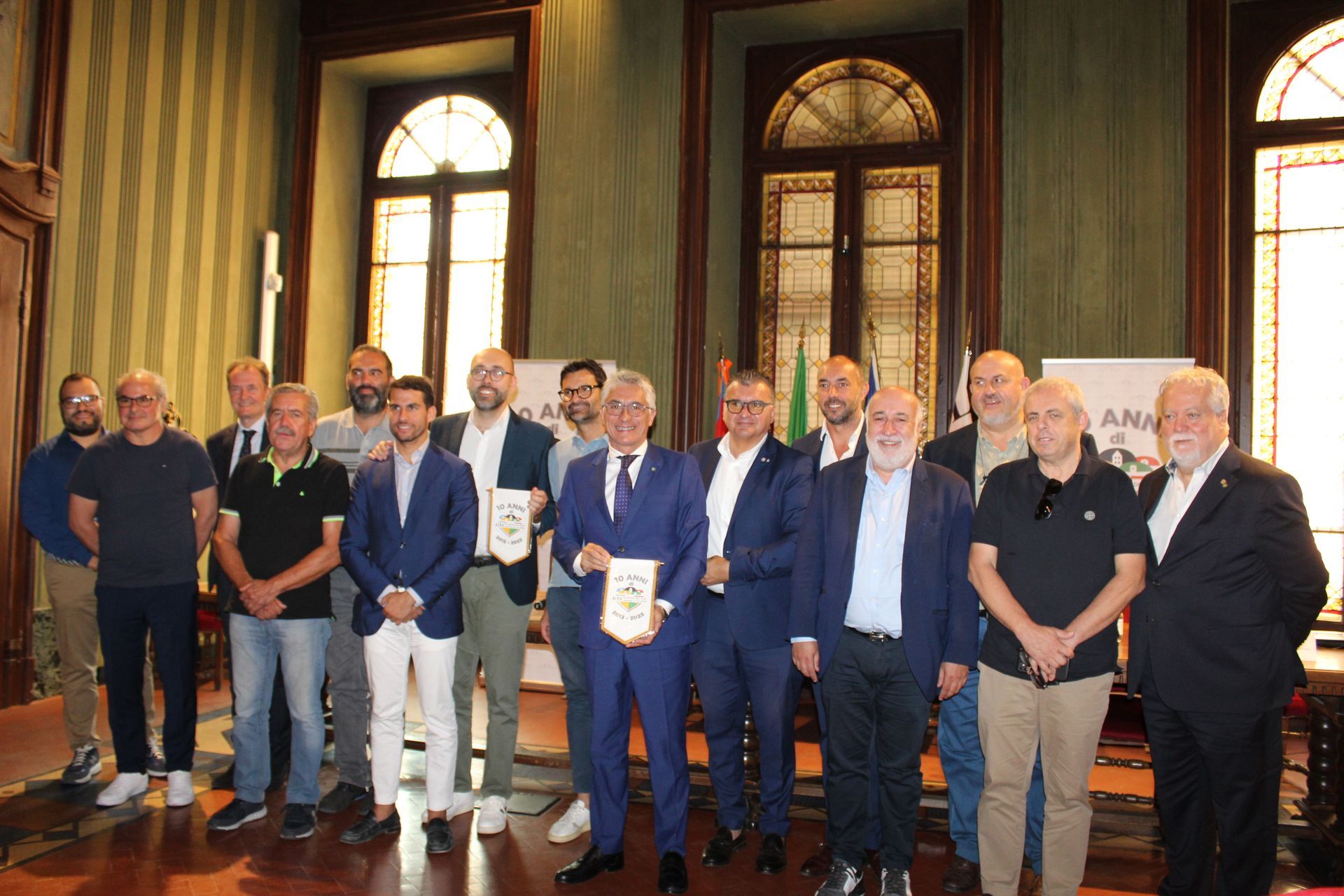 Gruppo di uomini in una stanza con premi in mano. Alcuni indossano abiti eleganti. Finestre in legno e decorazioni sullo sfondo.