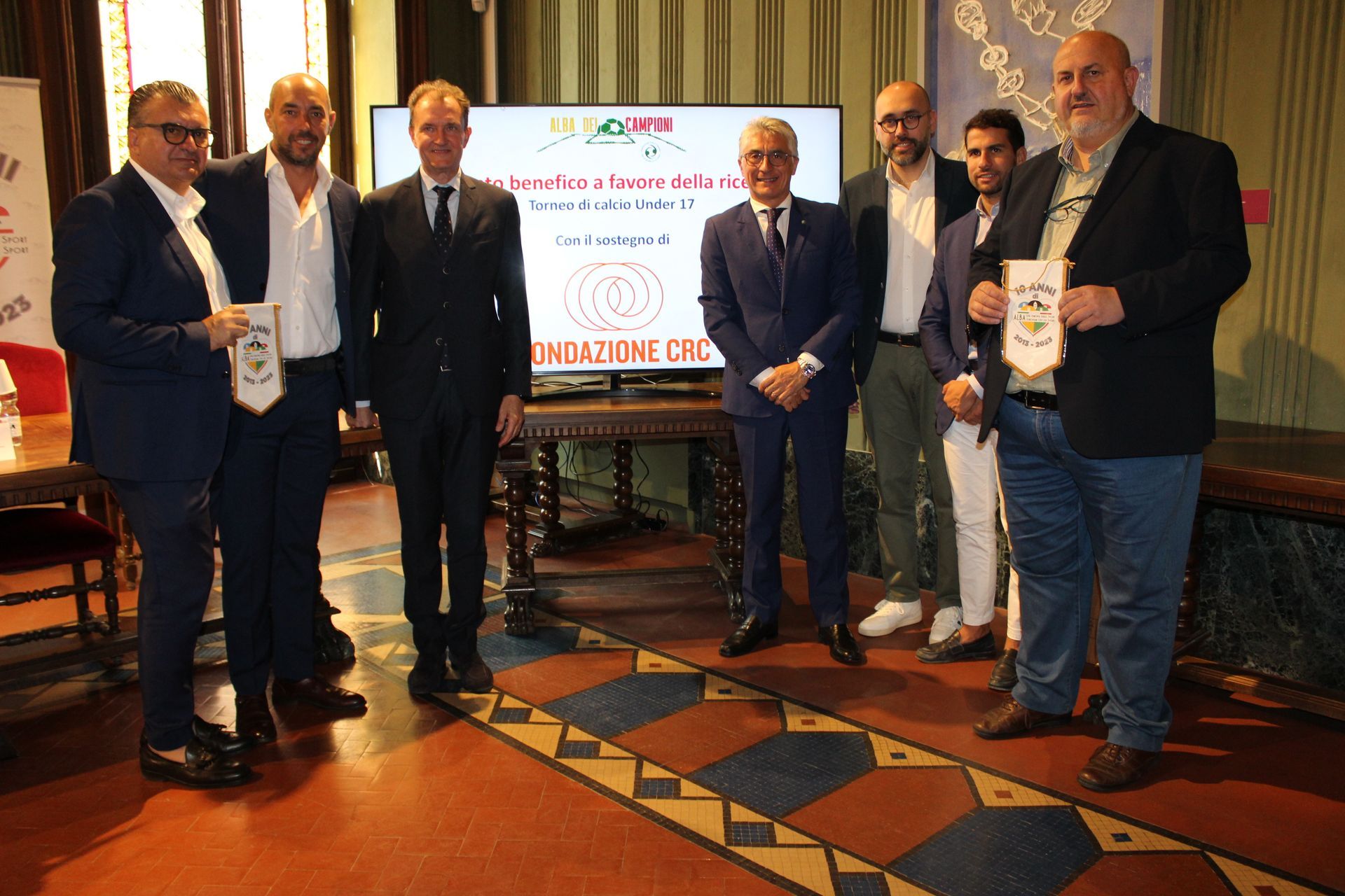 Un gruppo di uomini in giacca e cravatta, con in mano dei gagliardetti, si è riunito in una stanza dotata di uno schermo per le presentazioni.