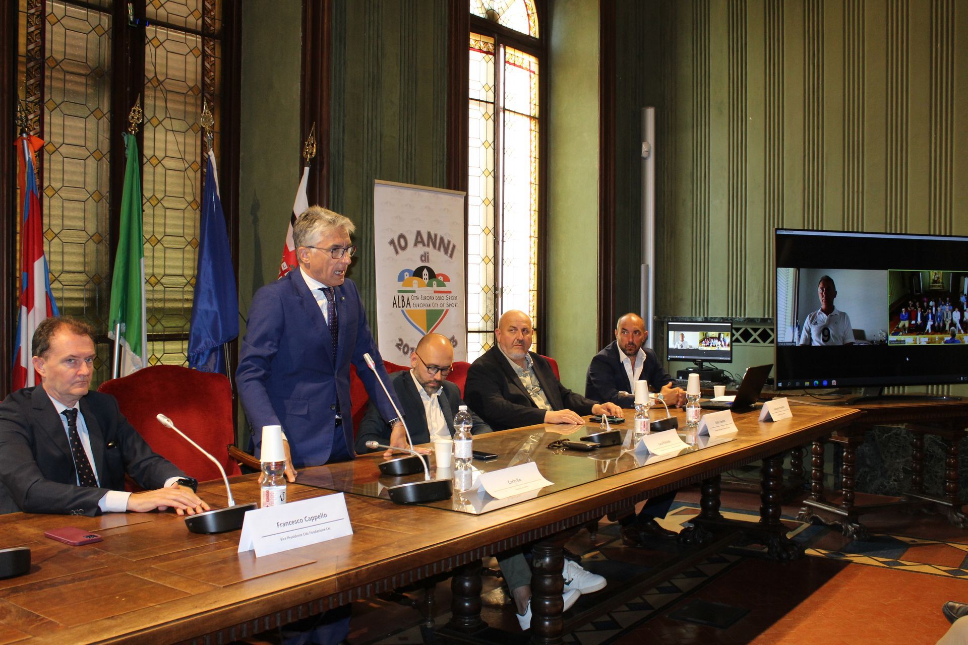 Persone sedute a un tavolo da conferenza, una che parla al microfono, l'altra su uno schermo tramite videochiamata.
