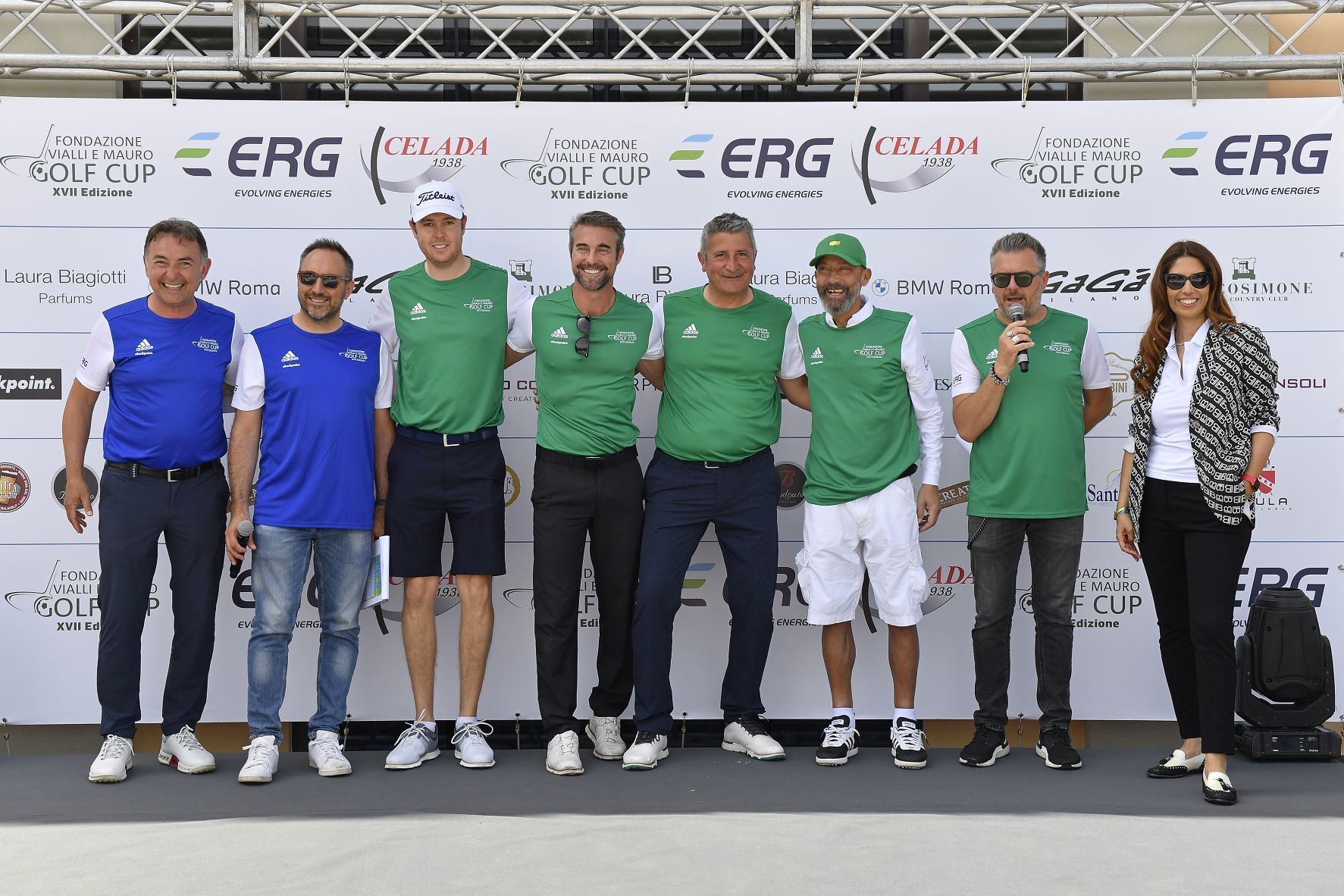 Un gruppo di nove persone in posa su un palco, alcune delle quali indossano gilet verdi. Il logo 