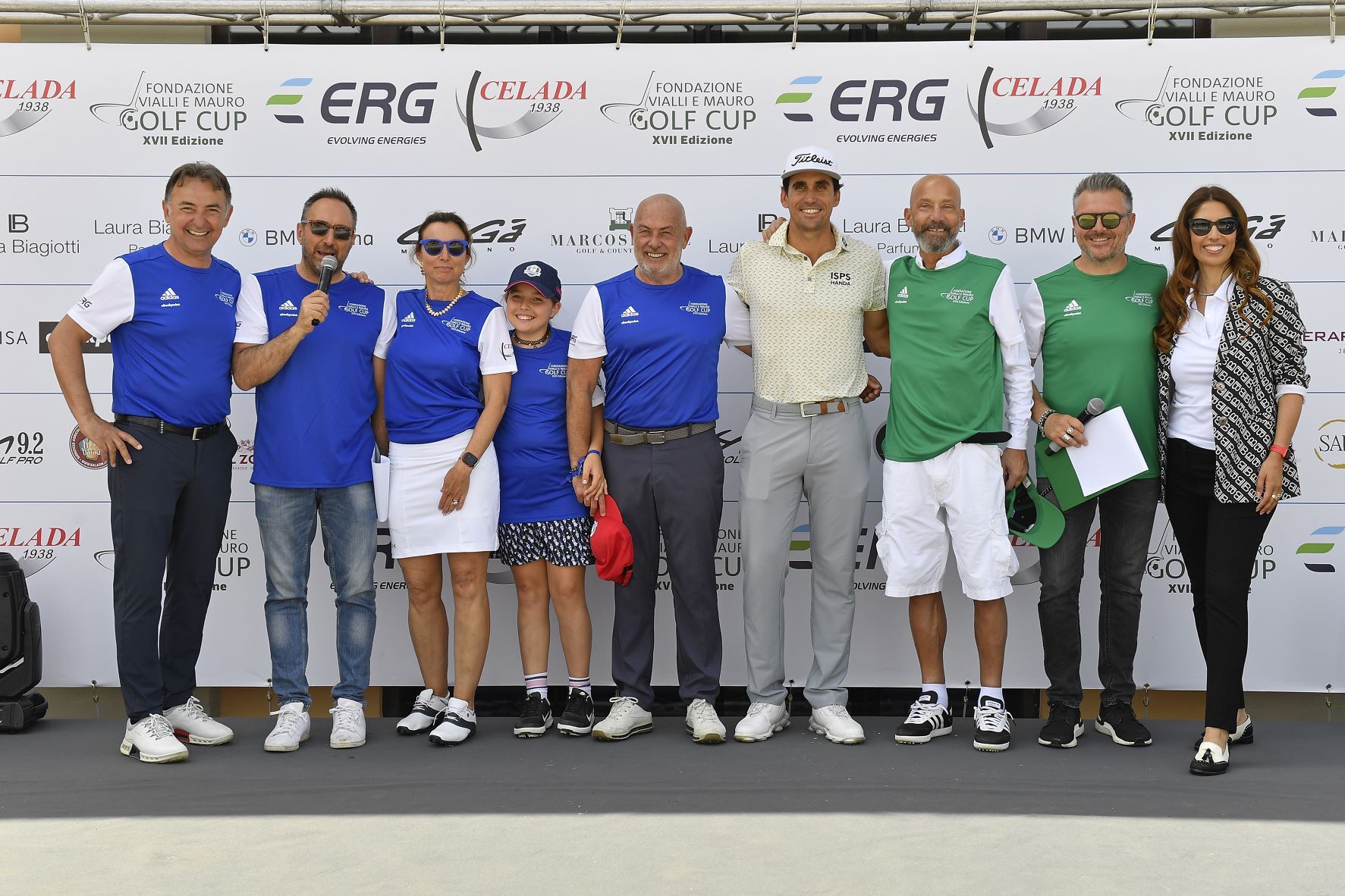 Gruppo di persone in posa per una foto a un evento di golf. Alcuni indossano divise blu o verdi e tengono in mano trofei e documenti.