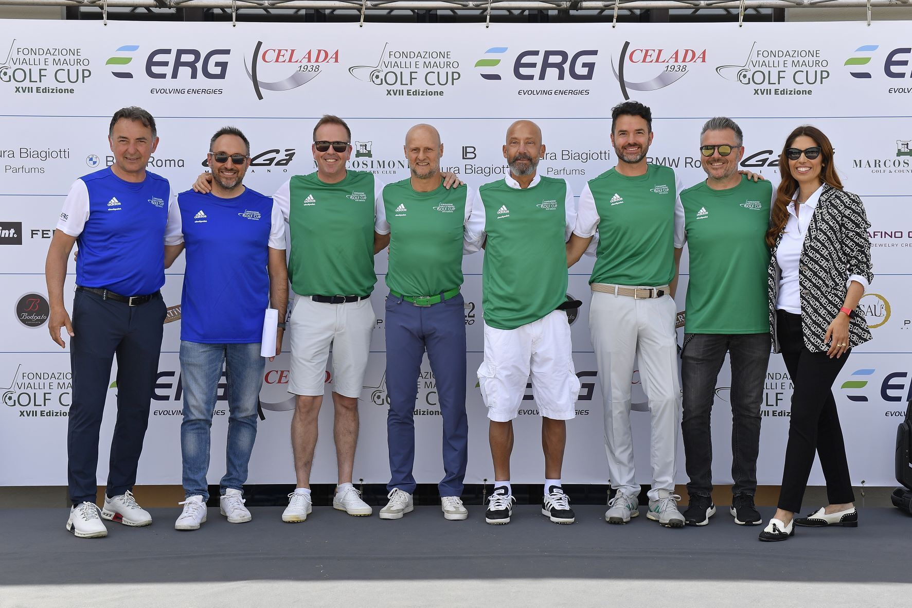 Gruppo di persone in posa per una foto a un evento di golf. La maggior parte indossa gilet verdi. Sullo sfondo c'è uno striscione.