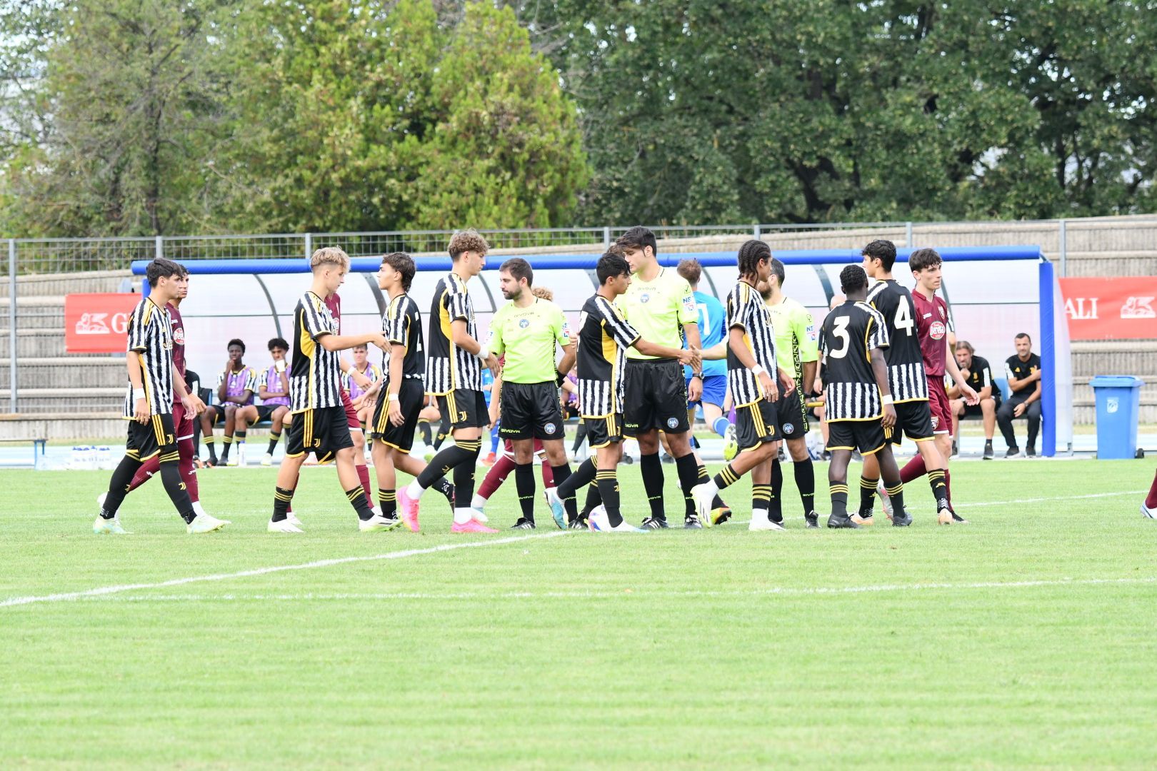 Giocatori di calcio con maglie a strisce bianche e nere e maglie color marrone che si stringono la mano su un campo verde.