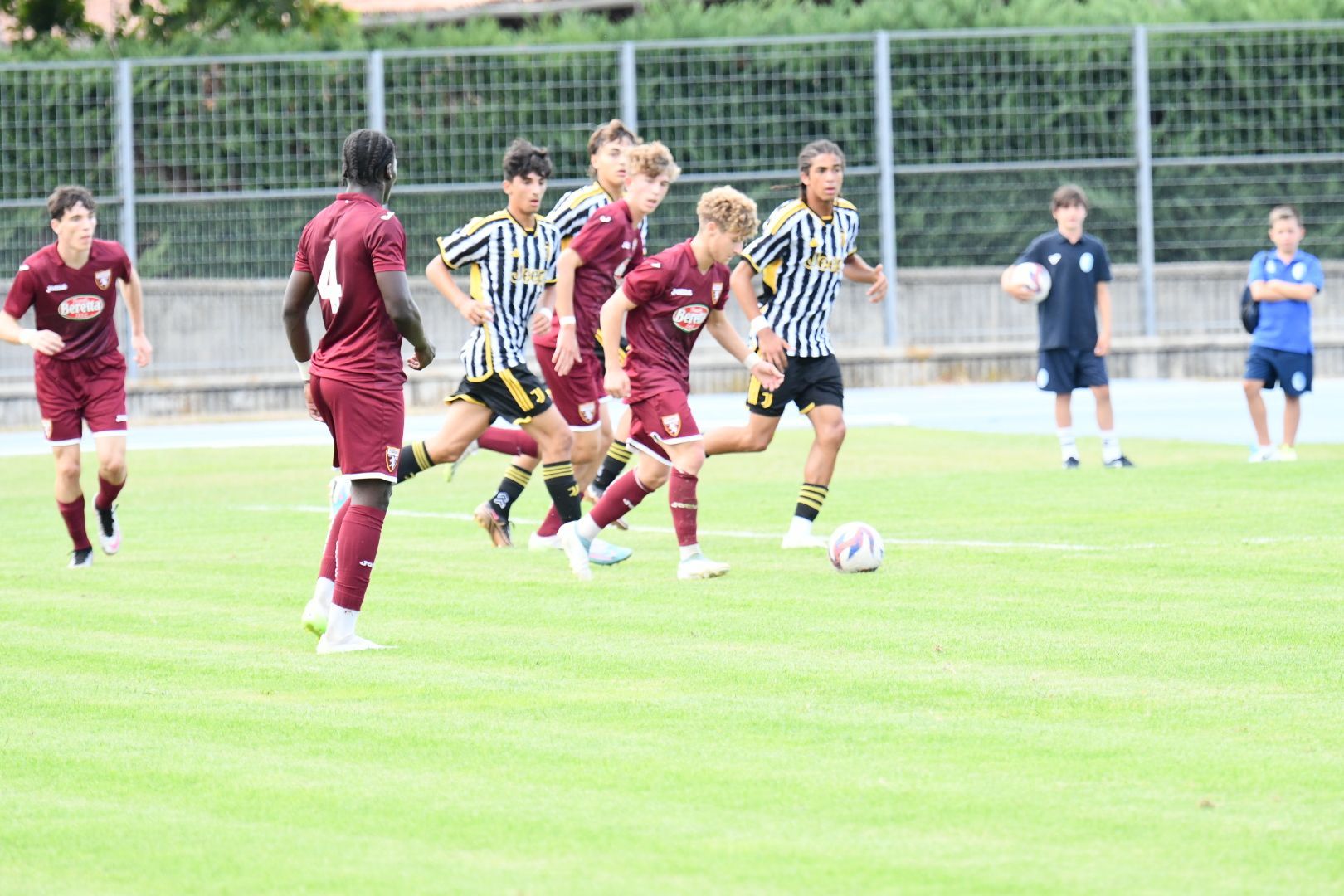 Giocatori di calcio in divise bordeaux e bianconere su un campo verde. Un giocatore calcia la palla.