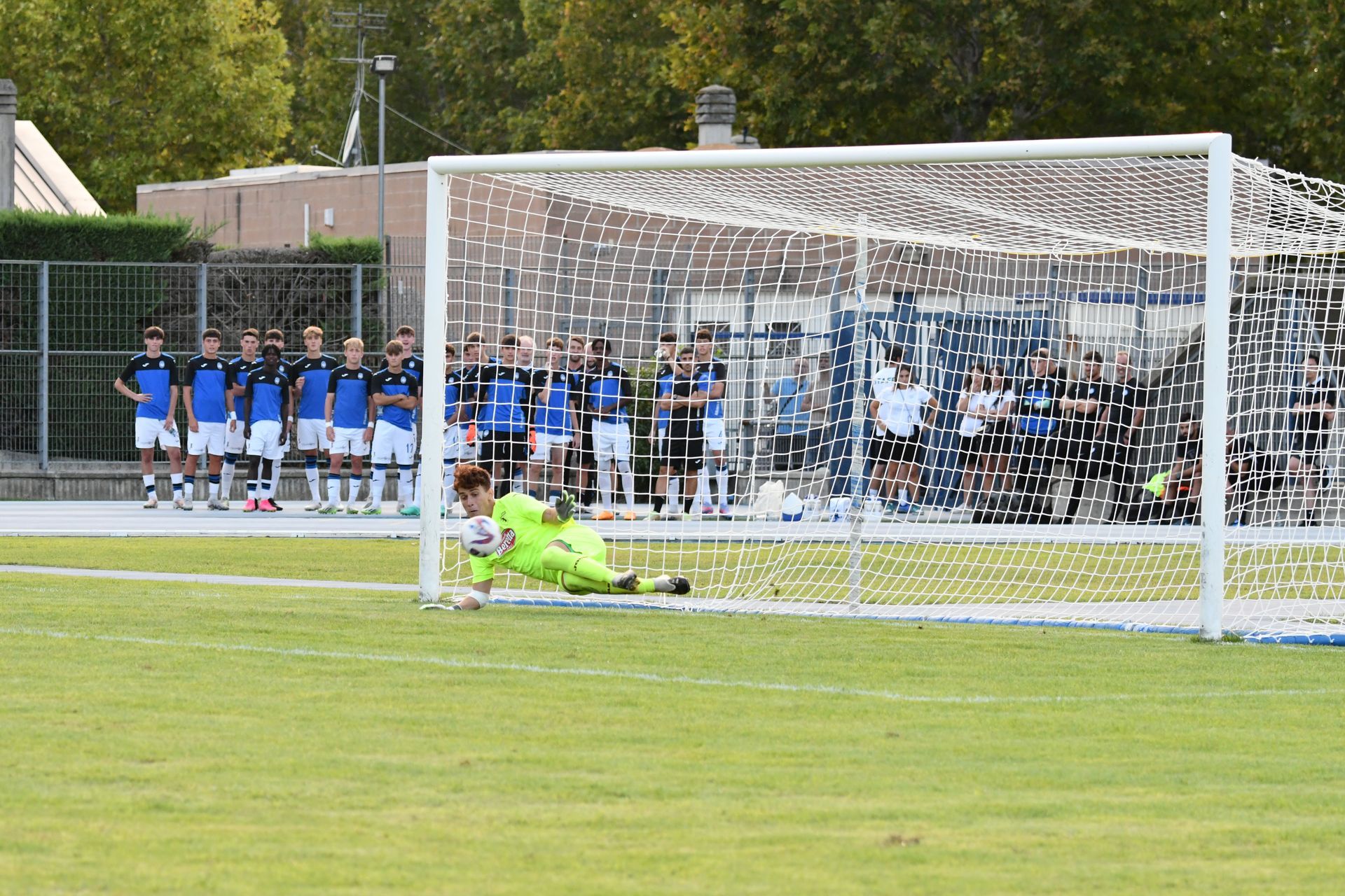 Un calciatore si tuffa per bloccare un calcio di rigore; la squadra in uniforme blu e bianca osserva dalla linea.