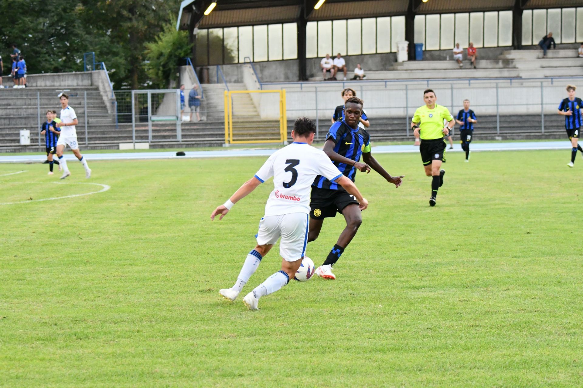 Giocatori di calcio in azione su un campo verde. Un giocatore vestito di nero cerca di dribblare un avversario vestito di bianco.